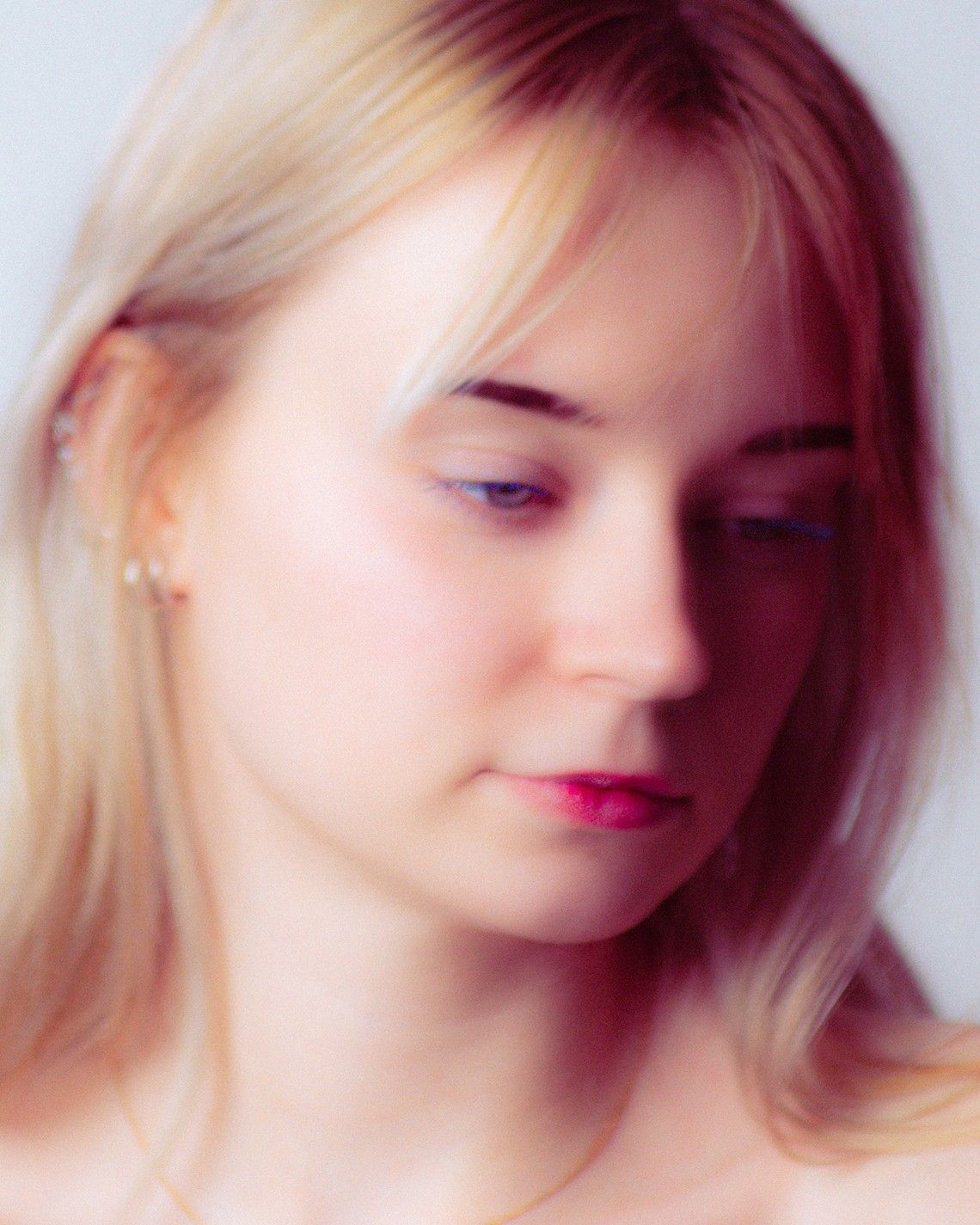 Soft-focus portrait of a young blonde woman with pink lips, looking down against a light background.