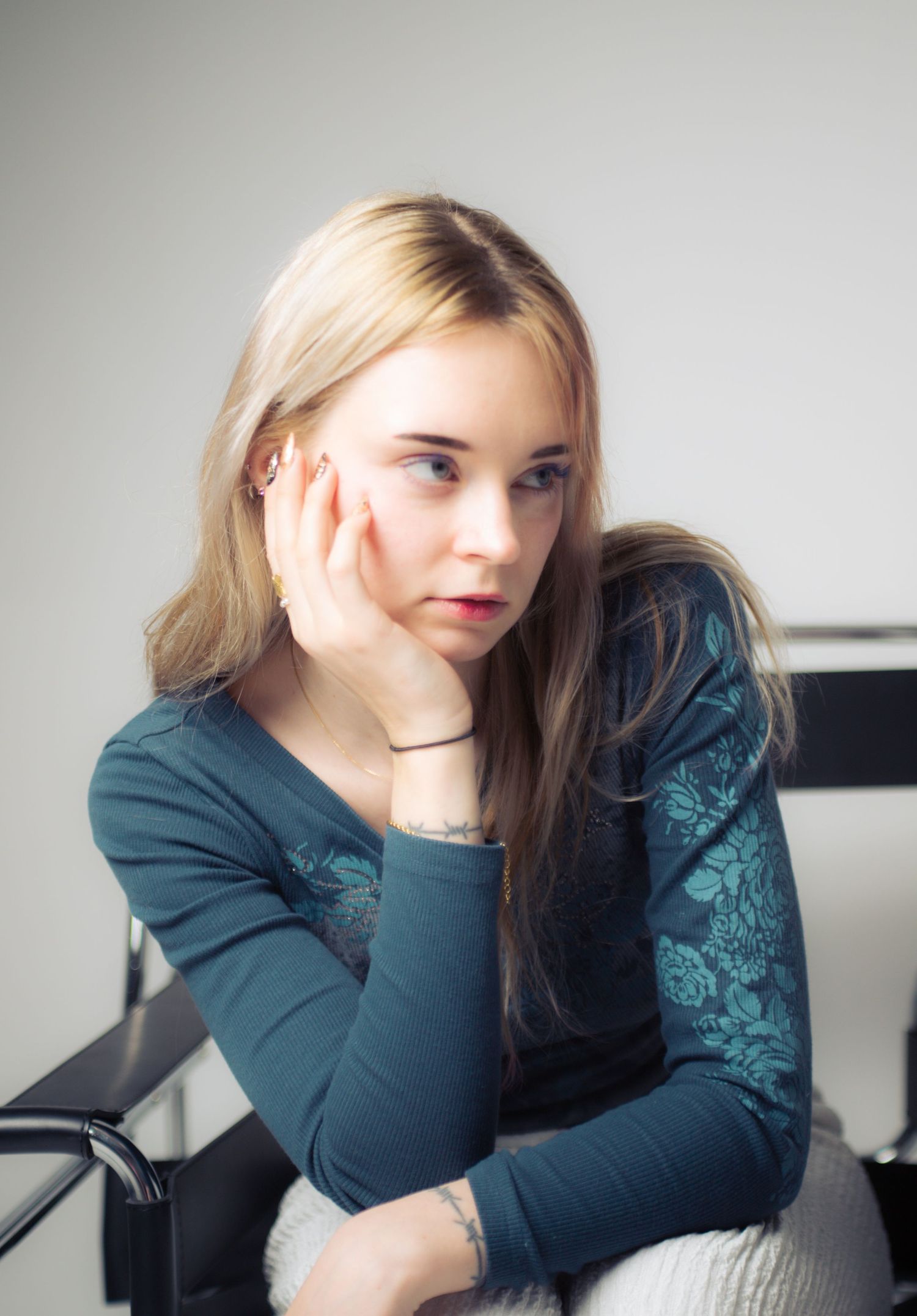 Young blonde woman in teal floral top sitting thoughtfully with hand on cheek in a modern interior.