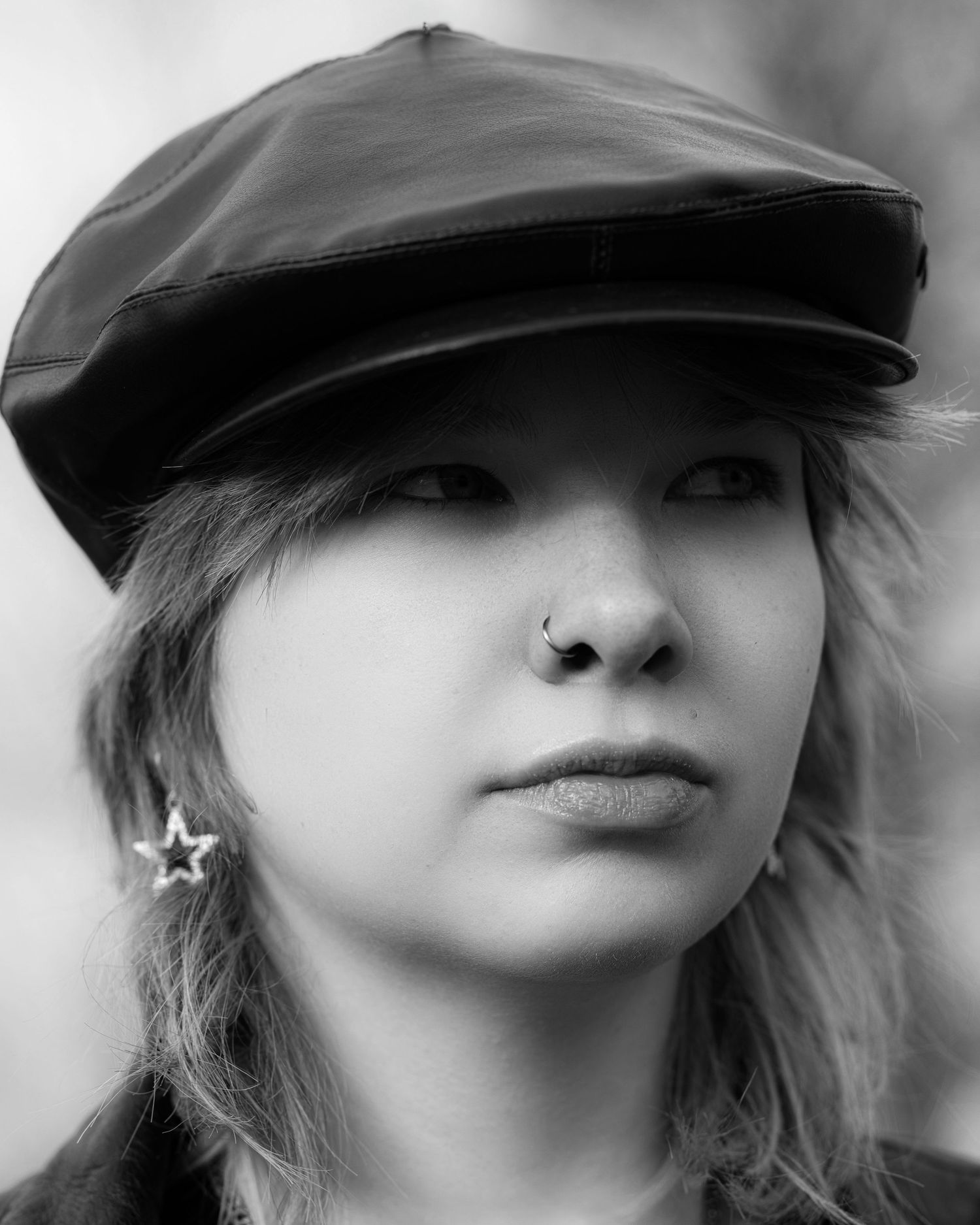 Black and white portrait of a young woman wearing a leather newsboy cap and star earring with a nose piercing.