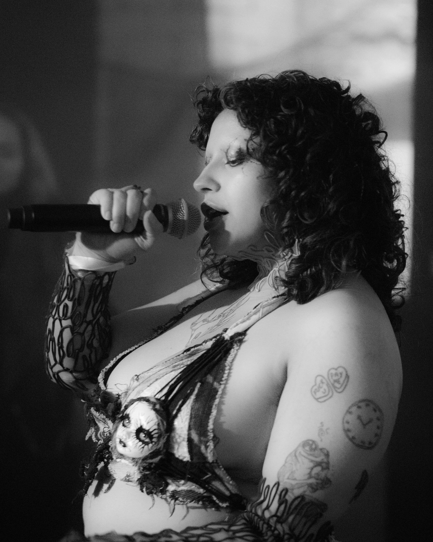 Black and white close-up of a tattooed female vocalist singing passionately into a microphone on stage.