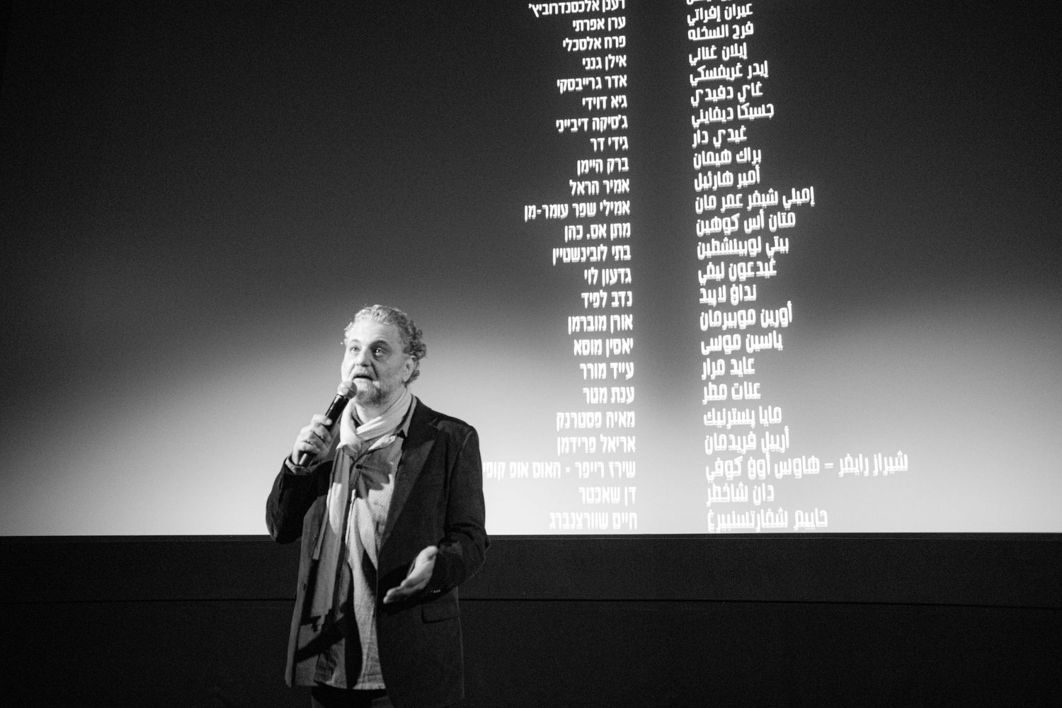 A black and white photo of a man speaking with a microphone in front of a screen displaying Hebrew text credits.