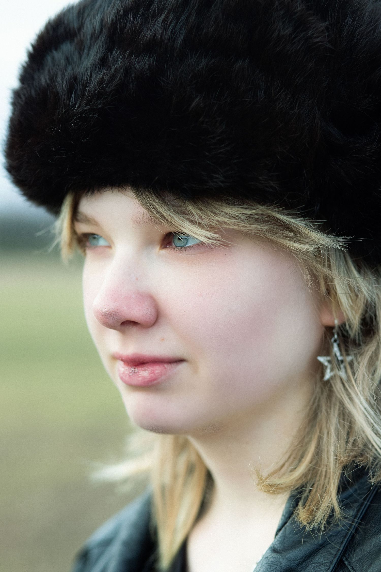 Young blonde woman wearing a black fur hat and leather jacket, with star earrings, gazing thoughtfully outdoors.