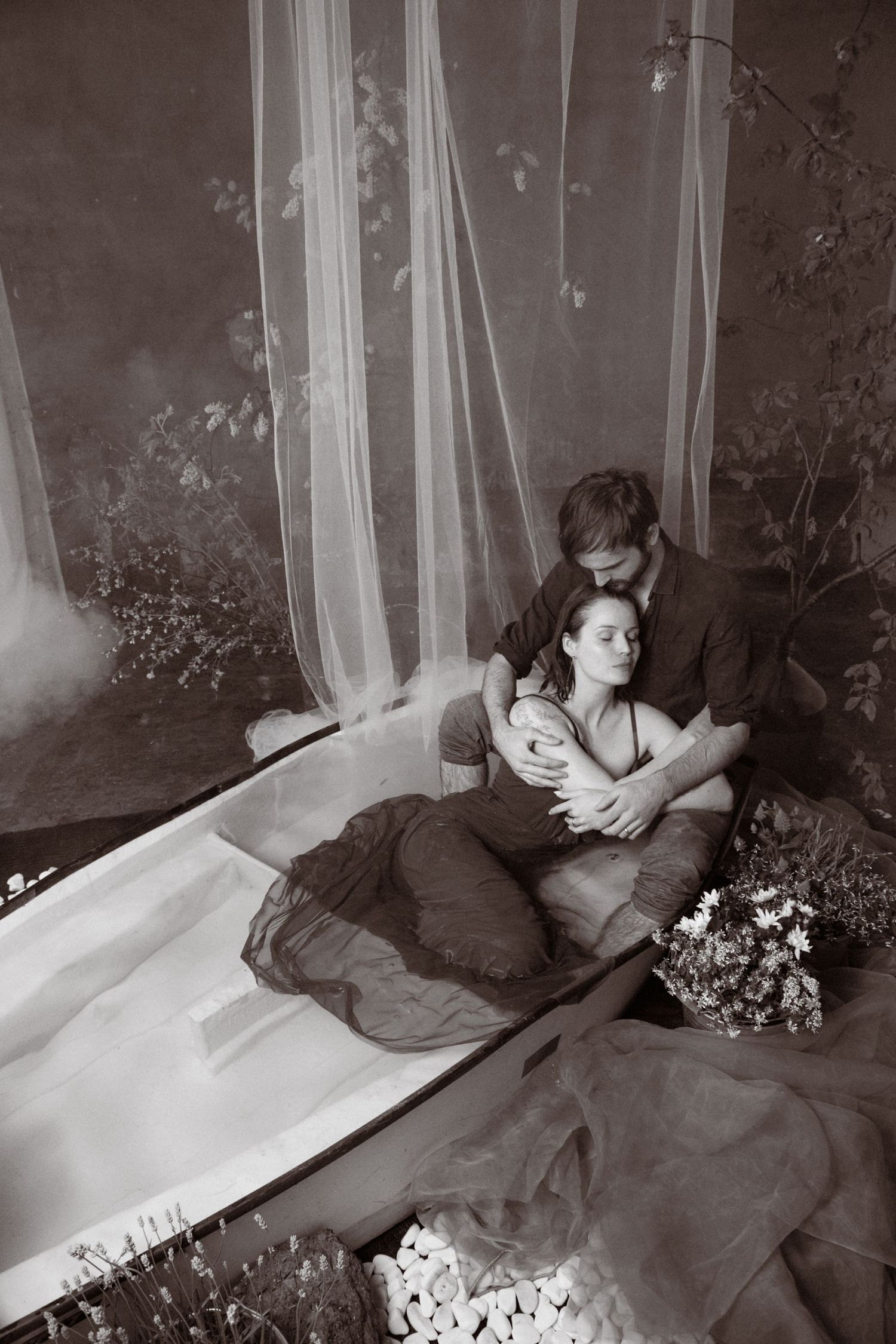 Black and white photo of a romantic couple embracing tenderly near a bathtub with sheer curtains and flowers.