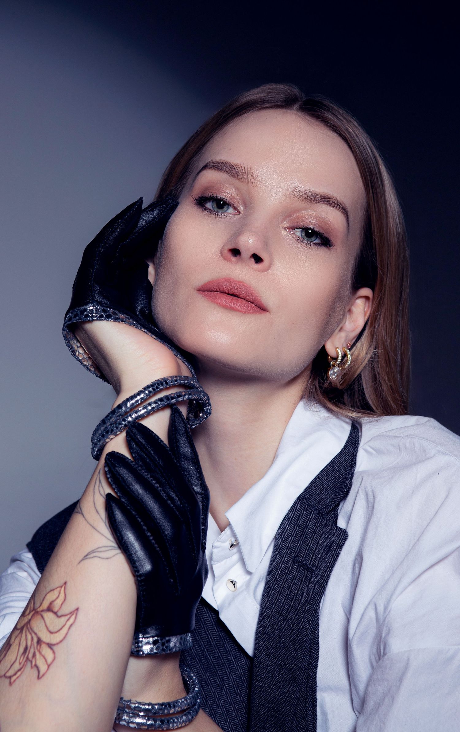 Woman in white shirt and black leather gloves poses with sparkling bracelets and a floral tattoo on her arm.