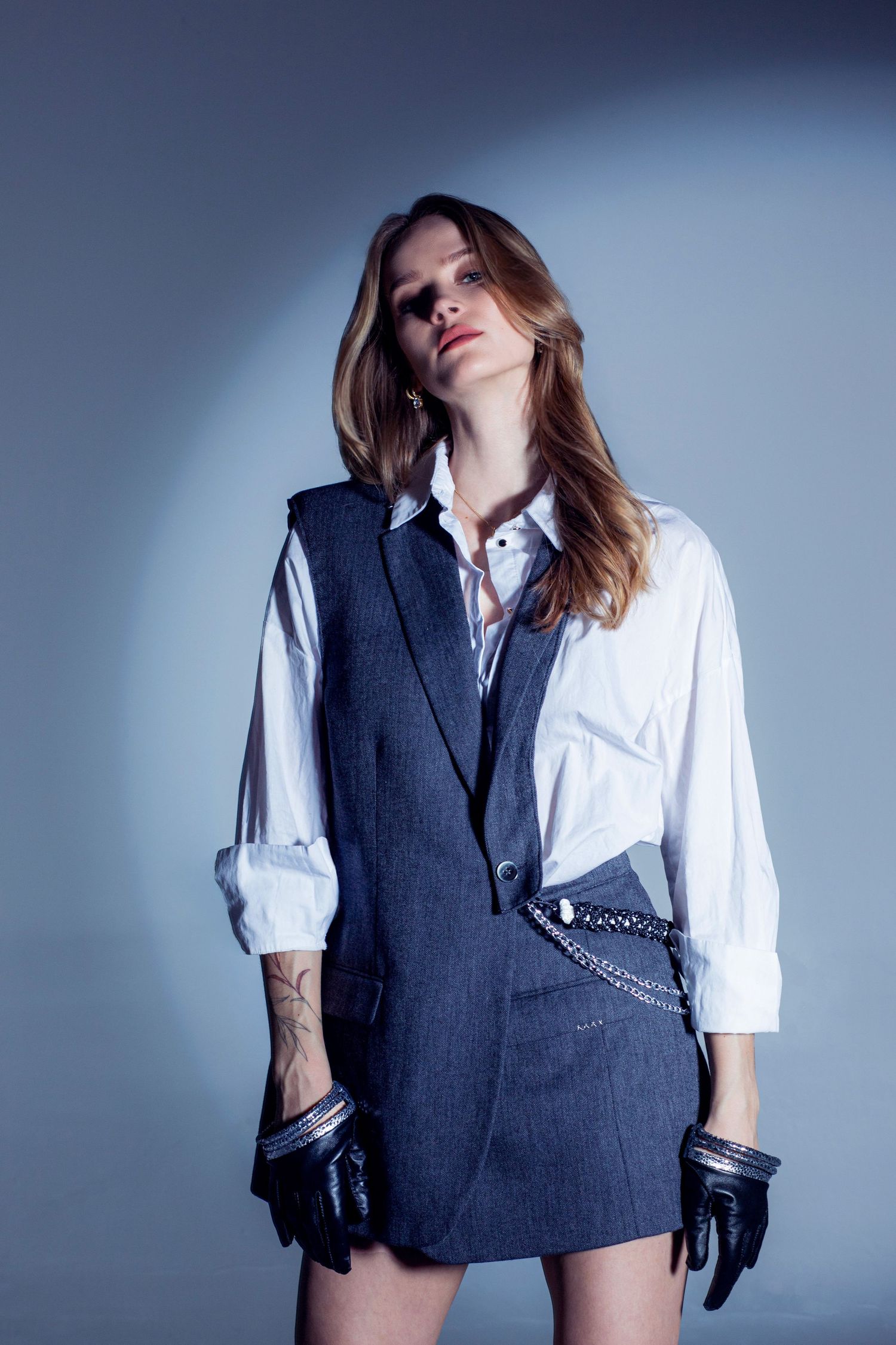 Woman in white shirt, denim vest, chain belt, and black leather gloves poses against a blue-gray studio background.