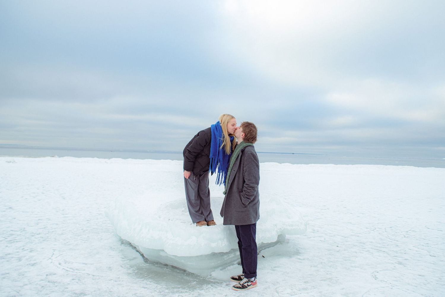 Couple sharing a romantic kiss on a frozen icy lake shore in winter, wearing cozy scarves and jackets.