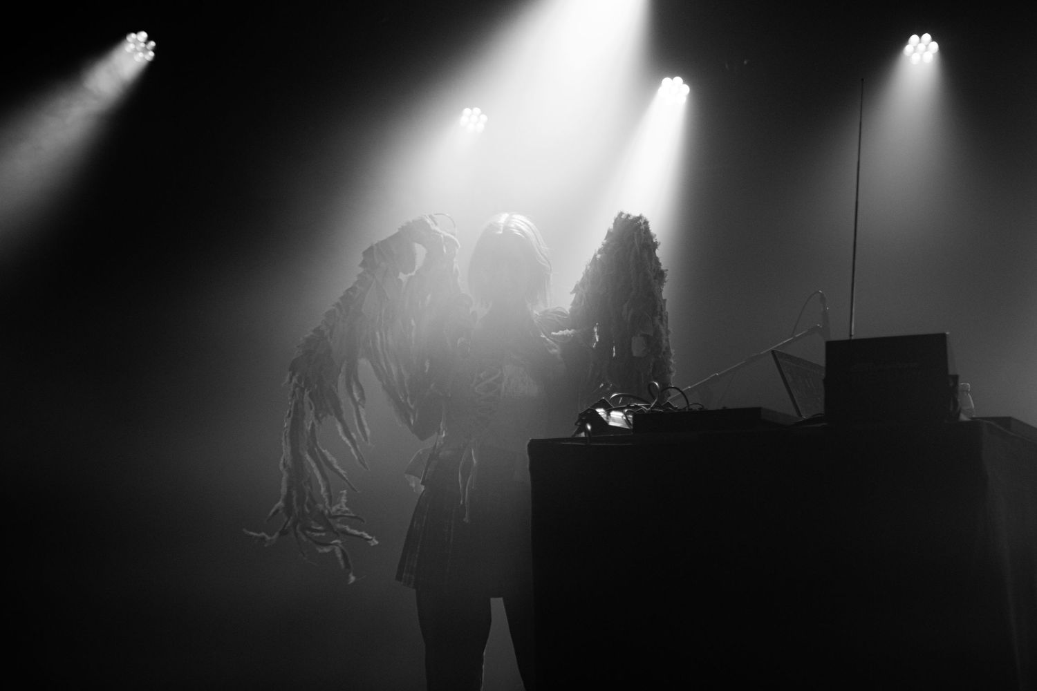 Dramatic black and white concert photo of performers on stage with spotlights and fog effects.