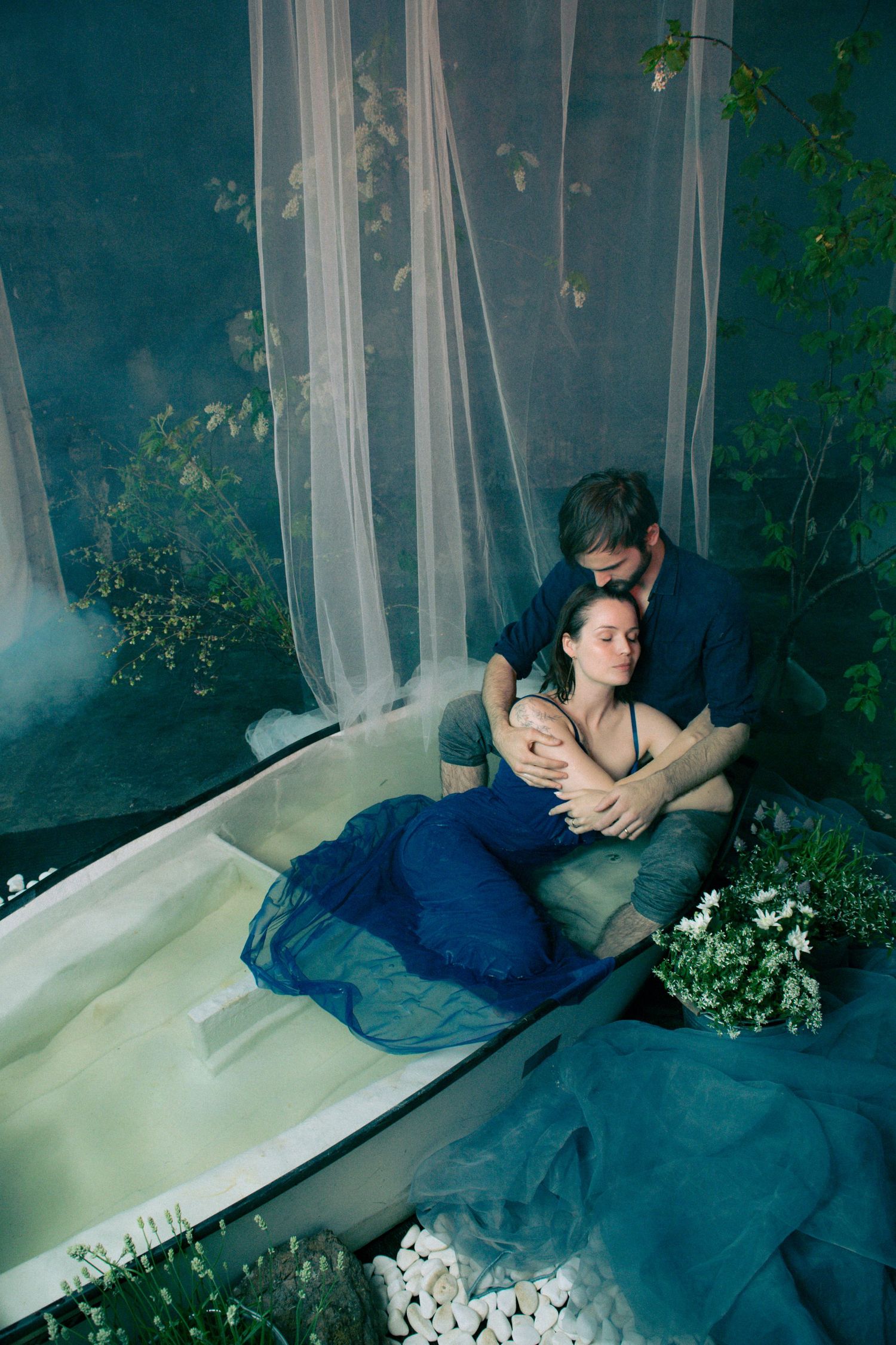 Couple embracing tenderly in a bathtub surrounded by lush greenery and sheer white curtains in moody blue tones.