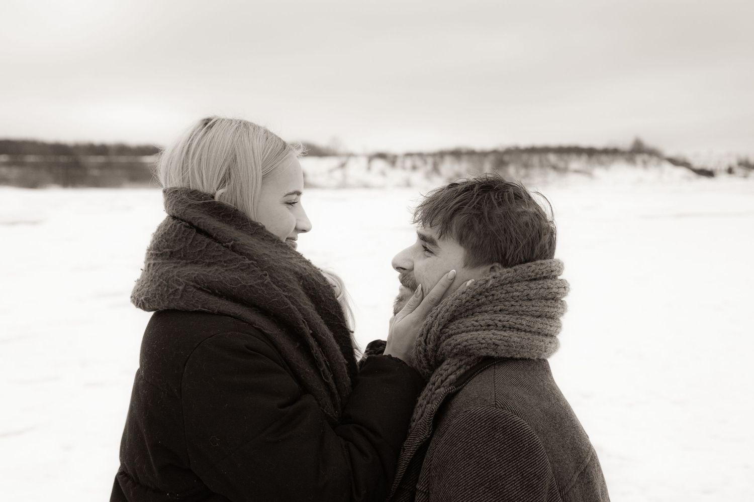 Couple sharing a romantic moment in snowy winter landscape, wearing scarves and dark coats, black and white photo.