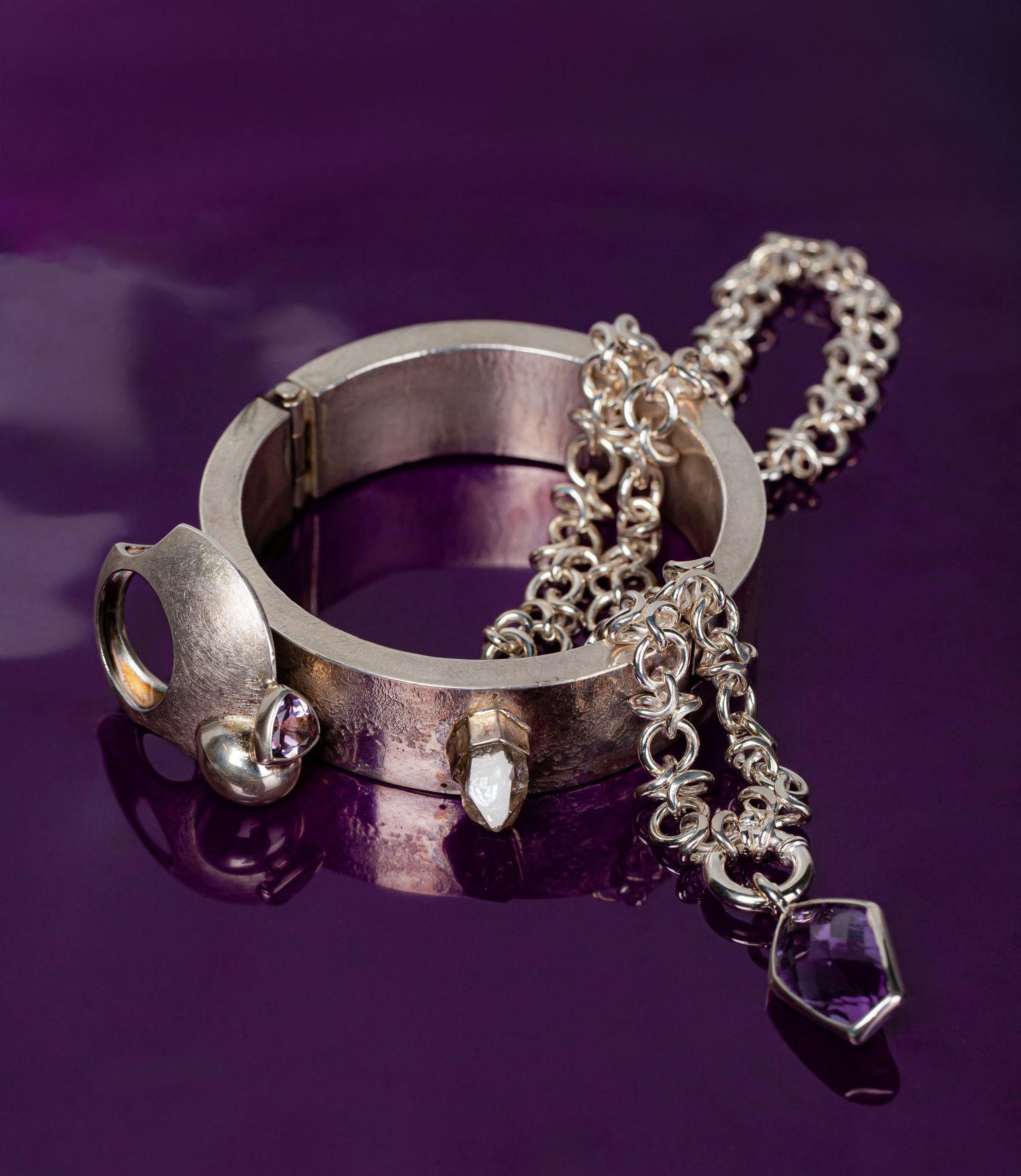 Silver bracelet with chain and amethyst crystal pendant on purple reflective surface.