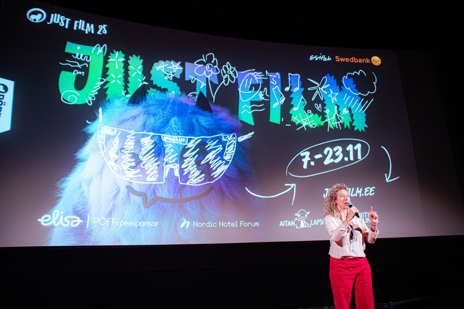 A woman presents on stage at the Just Film festival in front of a large promotional banner dated 7-23.11.
