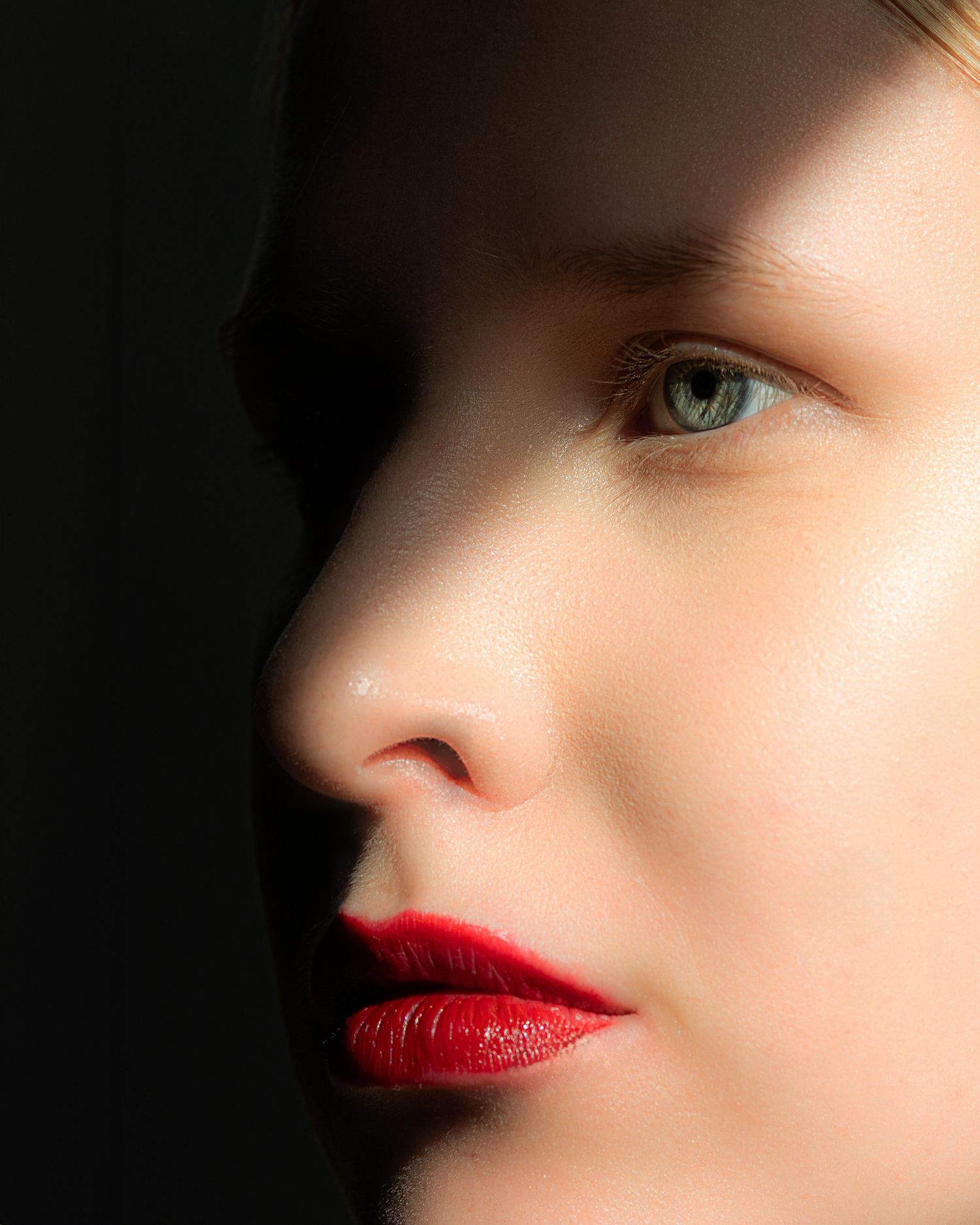 Close-up side profile of a woman with green eyes and bold red lips against a dark background.