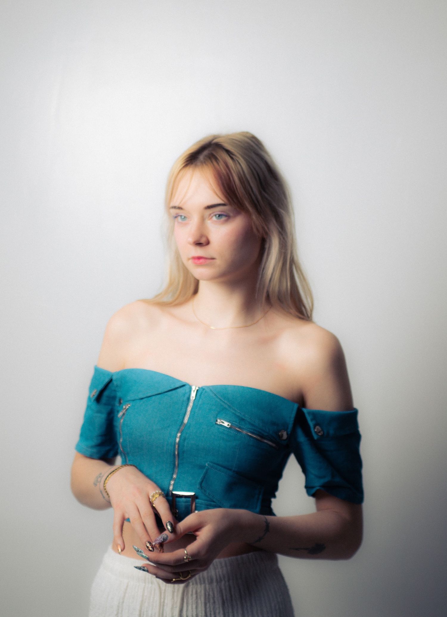 Young blonde woman in teal off-shoulder top holding a glass, posing against a light gray background.