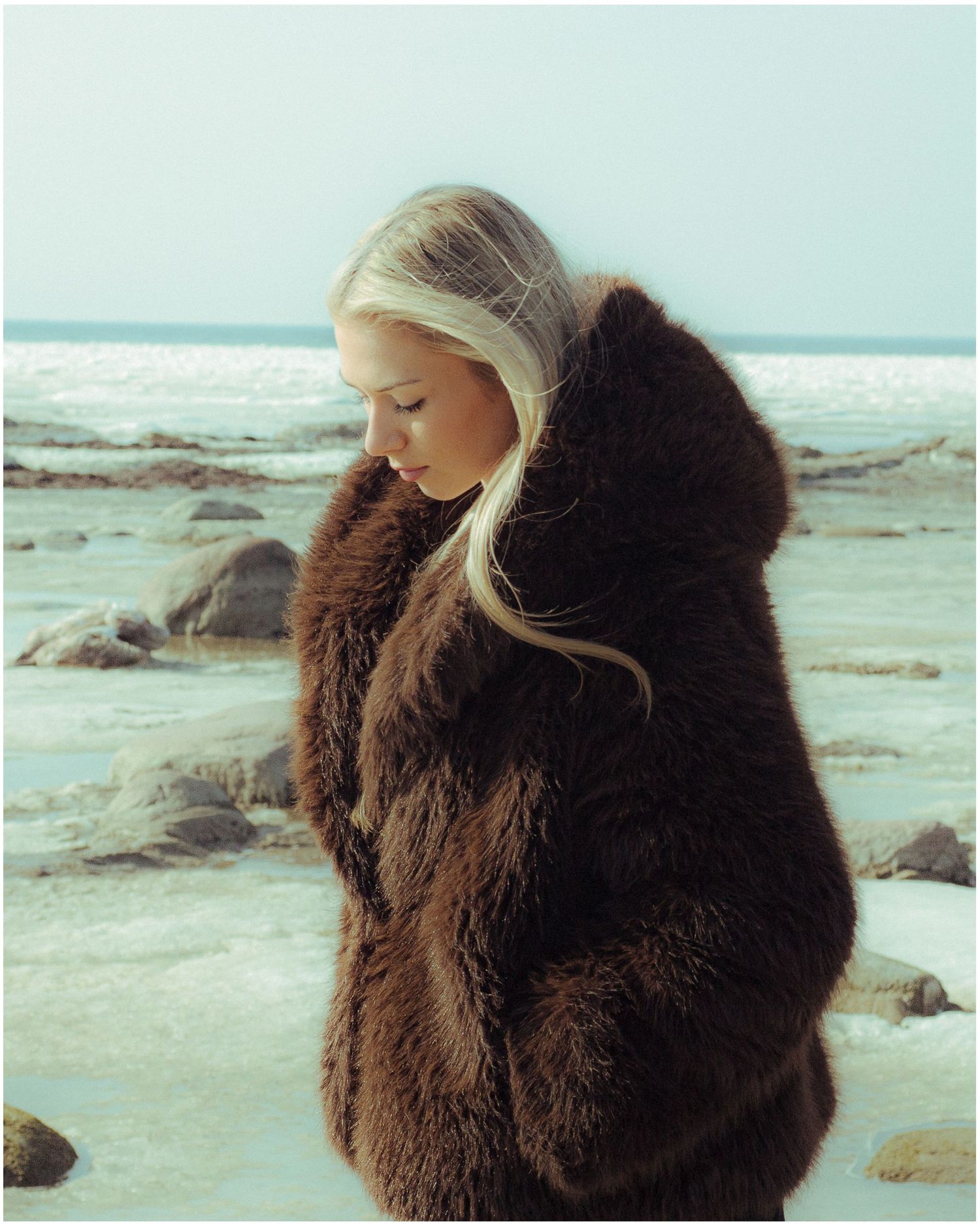 Blonde woman in oversized brown fur coat standing on rocky beach with ocean backdrop.