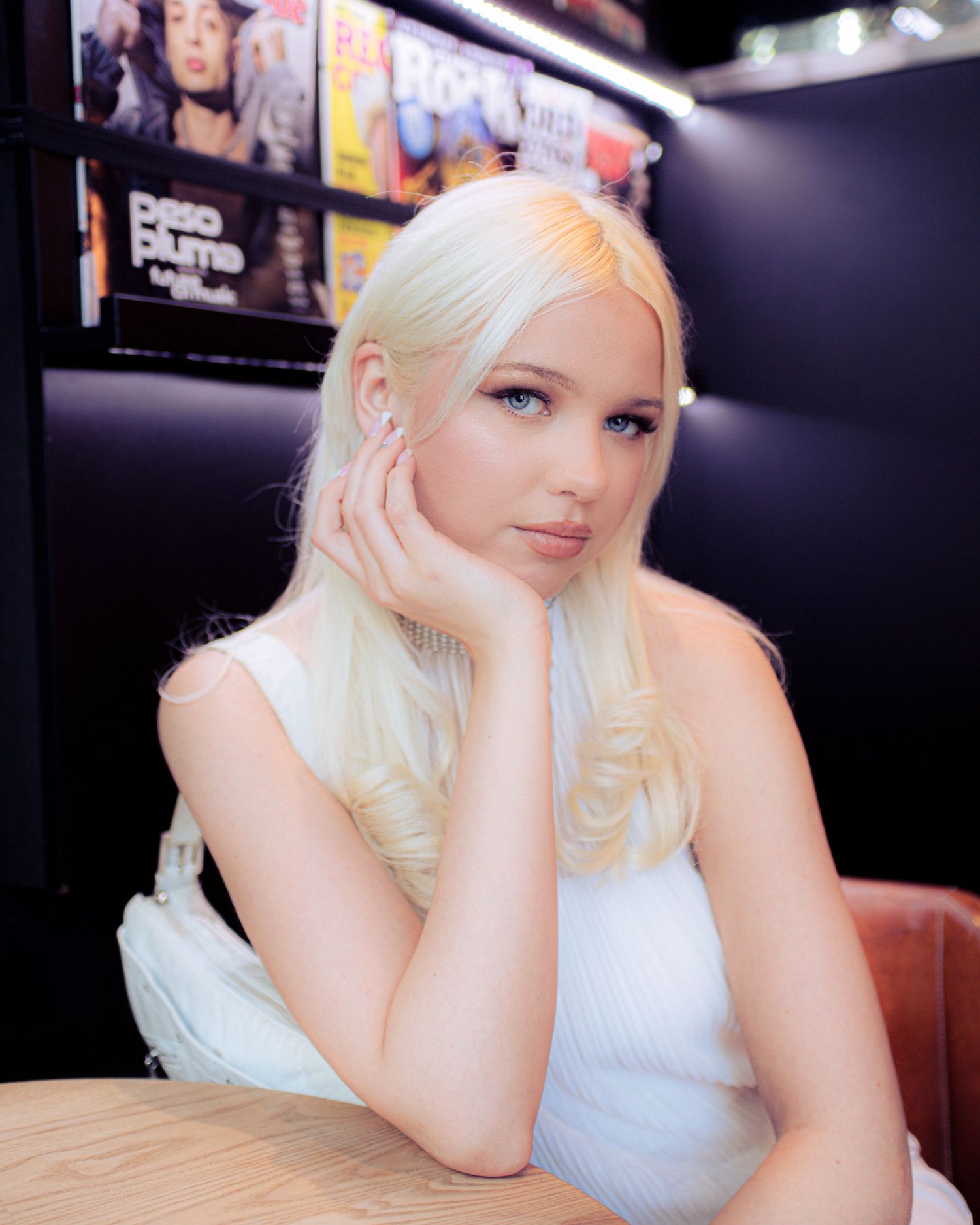 A young woman with long blonde hair and blue eyes poses at a table wearing a white sleeveless top.
