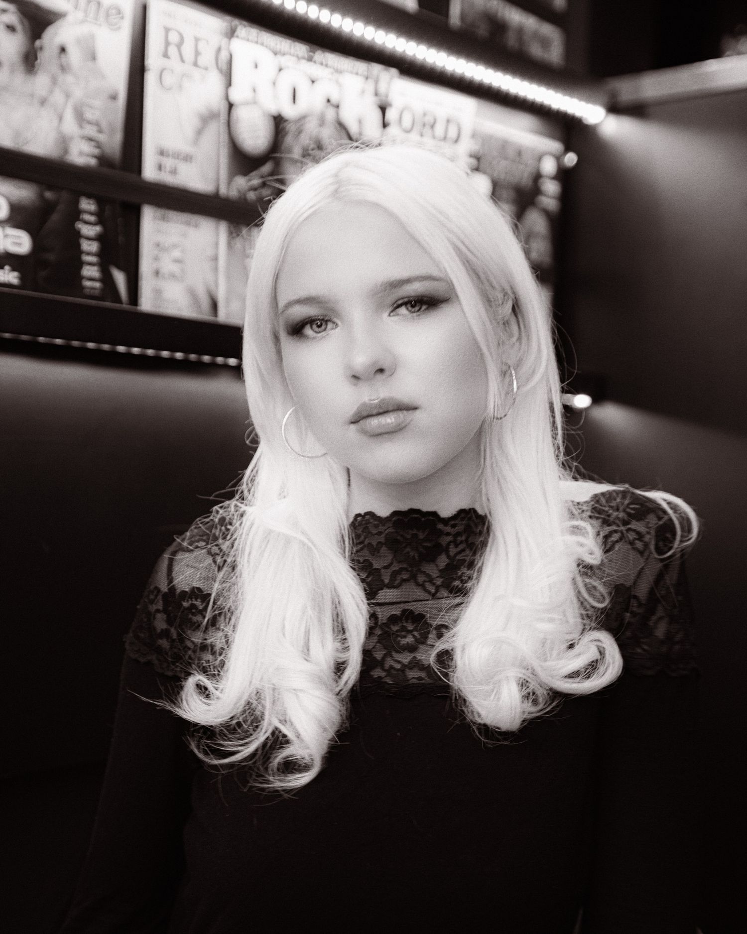 Black and white portrait of a young woman with long platinum blonde hair wearing a lace top in a record store.