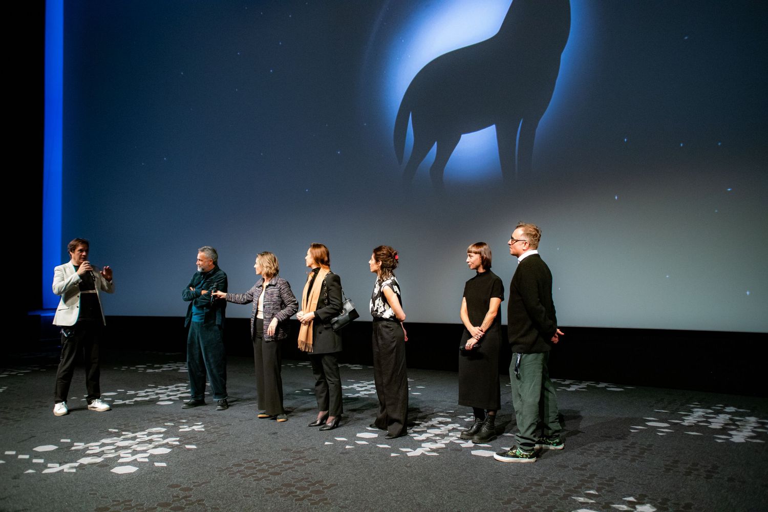 Group of people standing on stage at a film screening with a large wolf silhouette projected on the screen behind them.