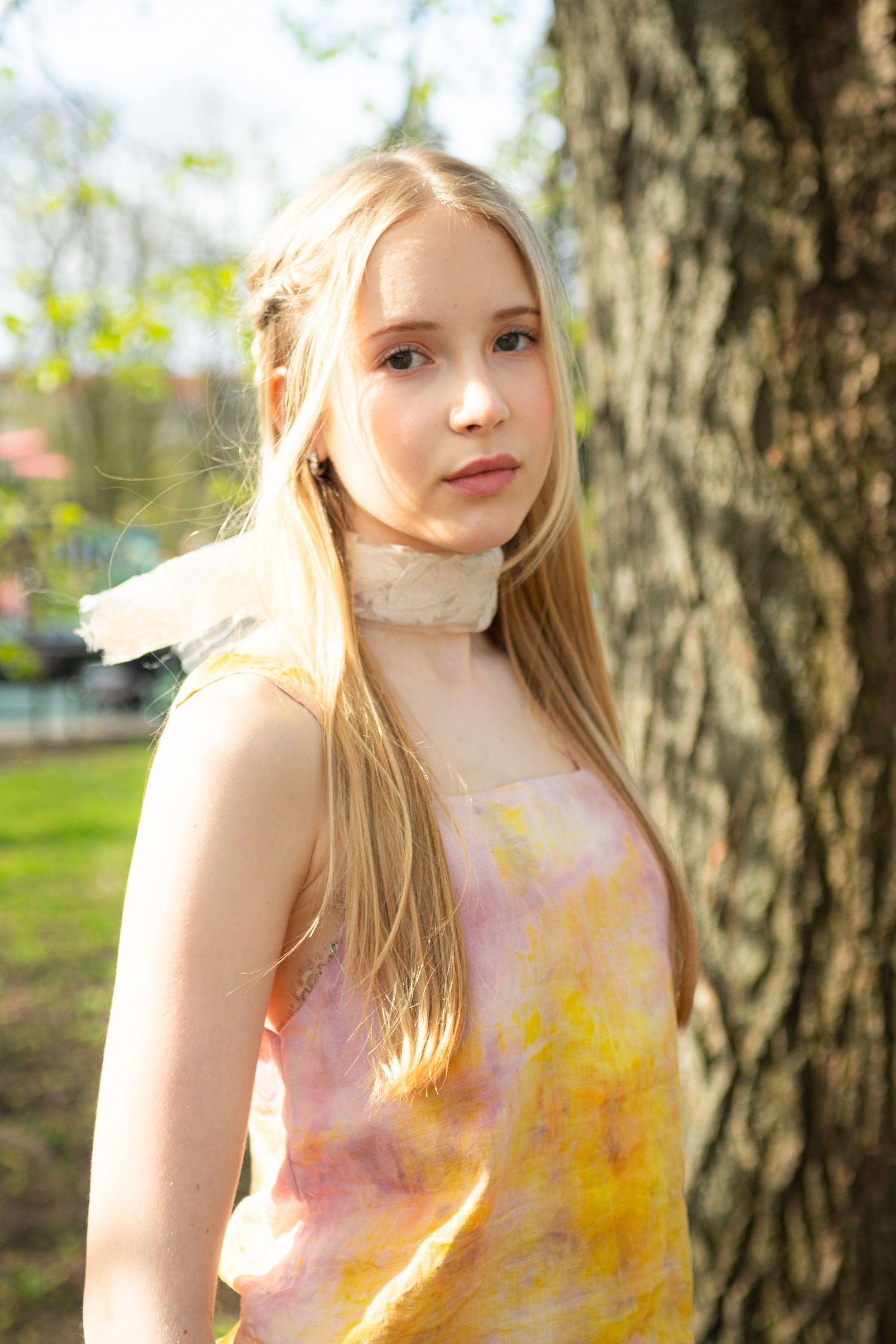 Young blonde woman in colorful tie-dye dress leaning against tree trunk in sunny park.