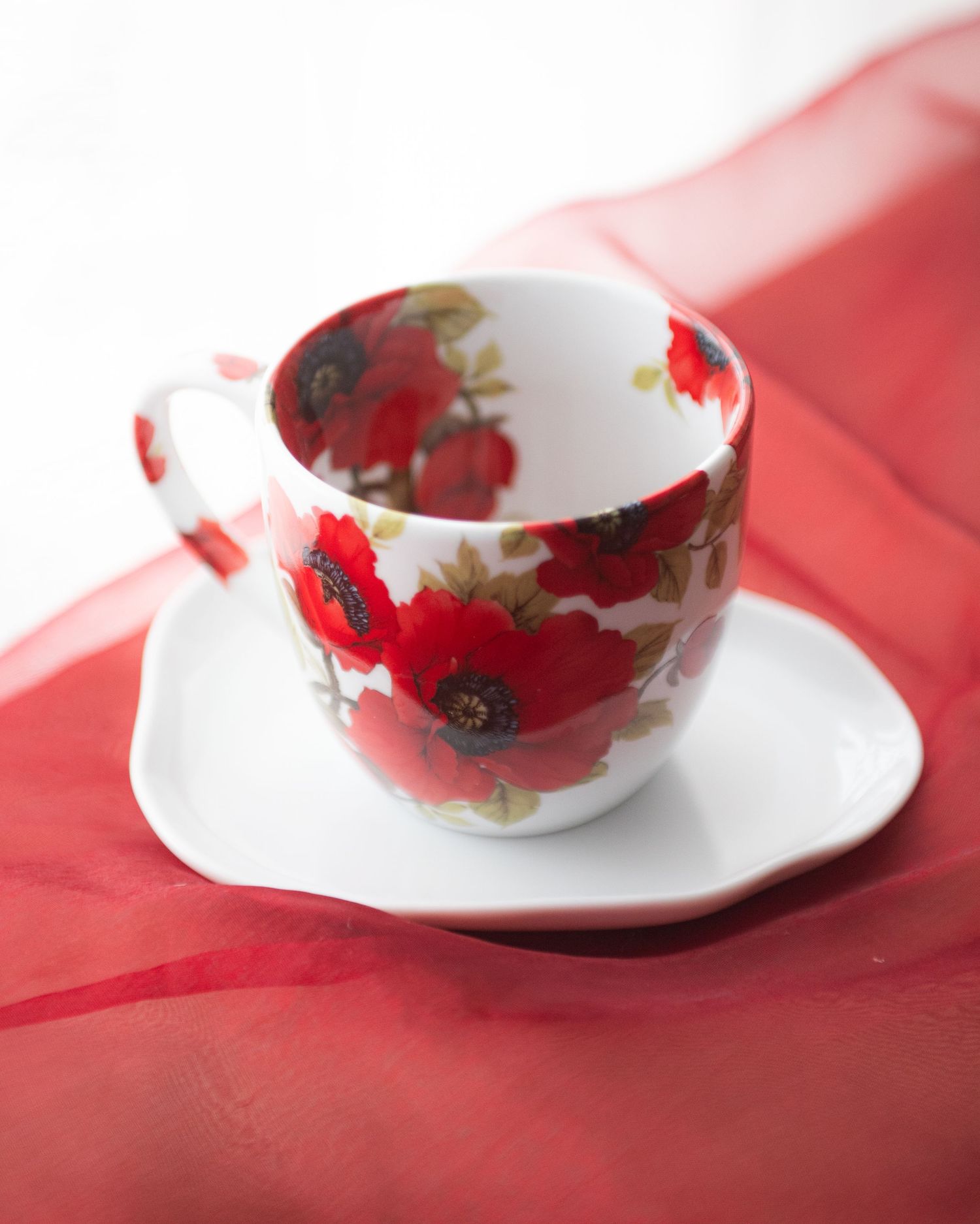 White porcelain teacup and saucer featuring vibrant red poppy floral design, placed on a red fabric surface.