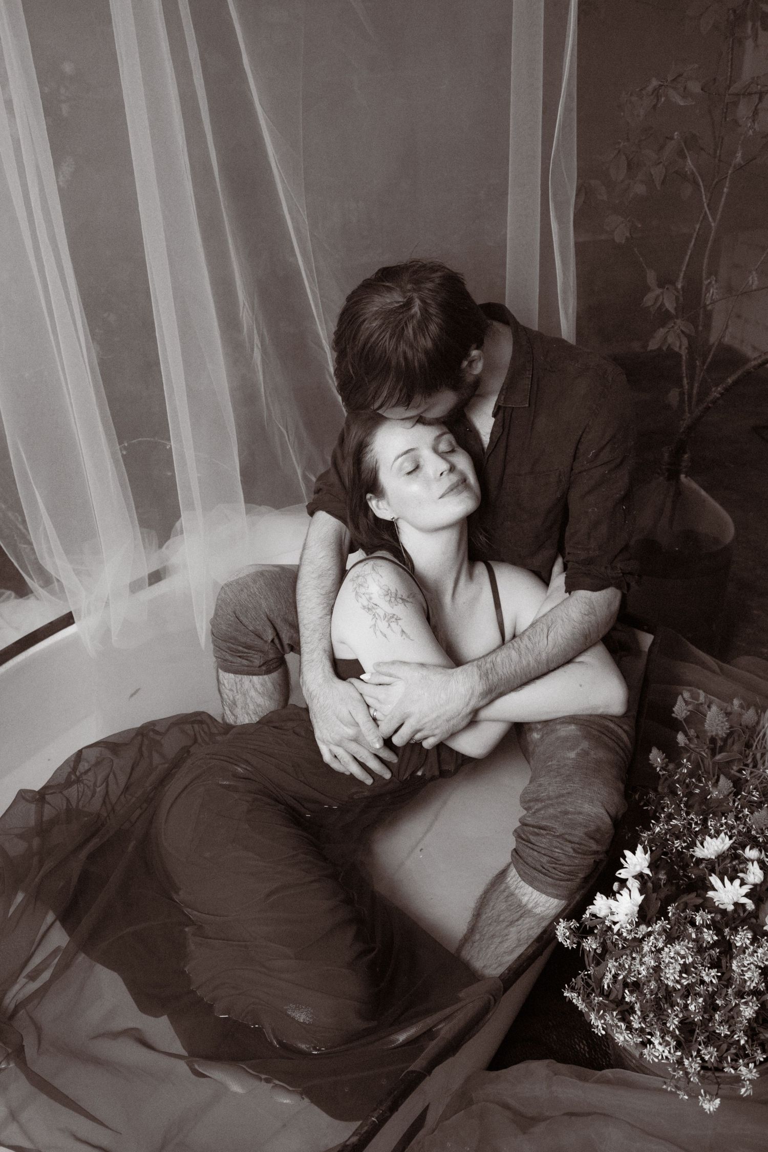 Black and white photo of a romantic couple embracing in a bathtub surrounded by sheer curtains and flowers.