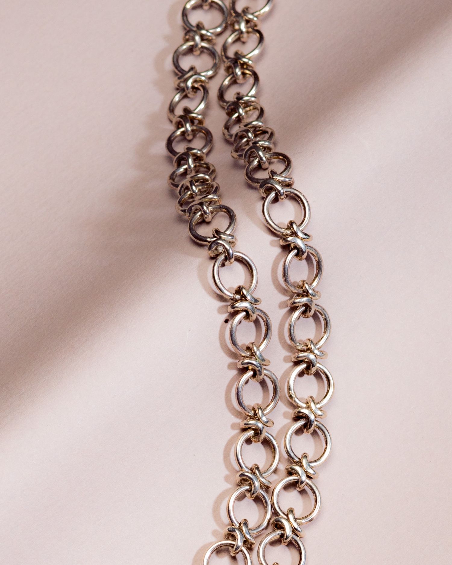 Close-up of a silver interlocking chain necklace with circular links, displayed on a soft pink fabric background.
