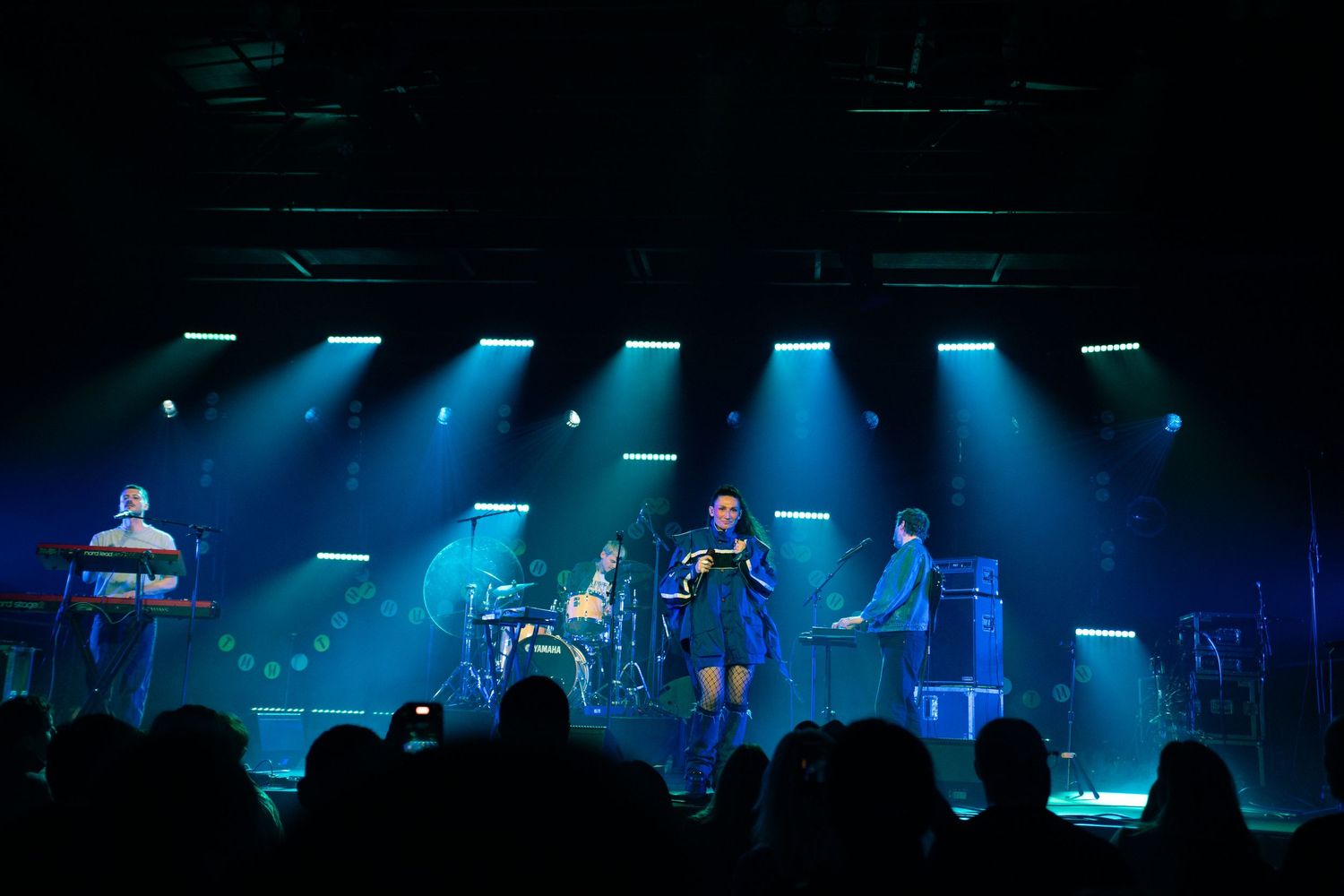 Band performing on a blue-green lit stage with silhouetted audience watching from below.