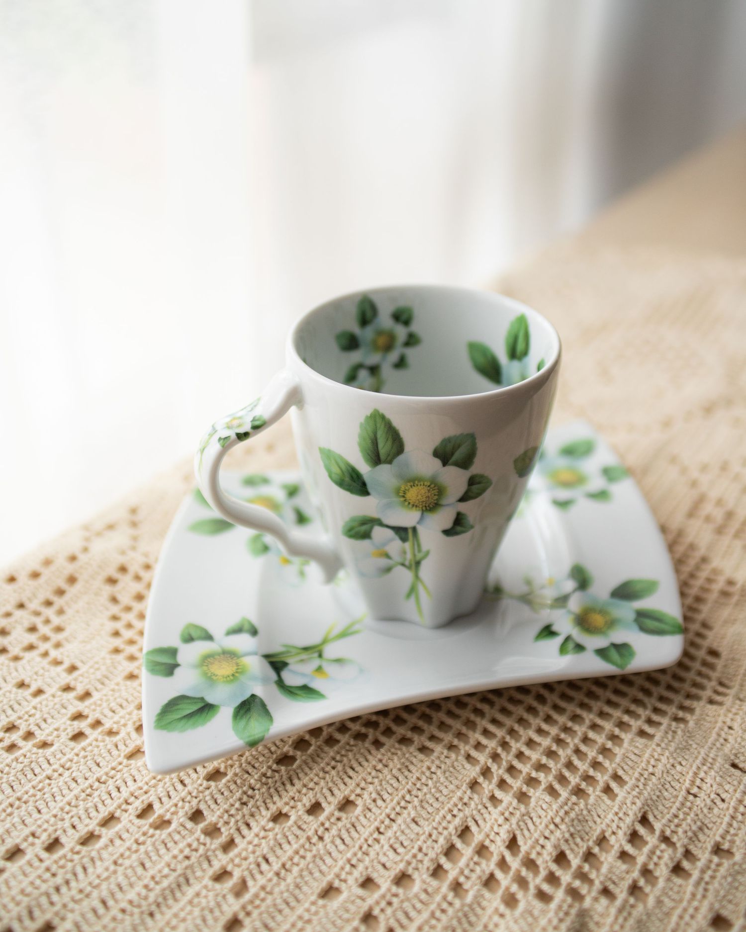 Elegant white porcelain teacup and saucer with green floral design on woven tablecloth near window.