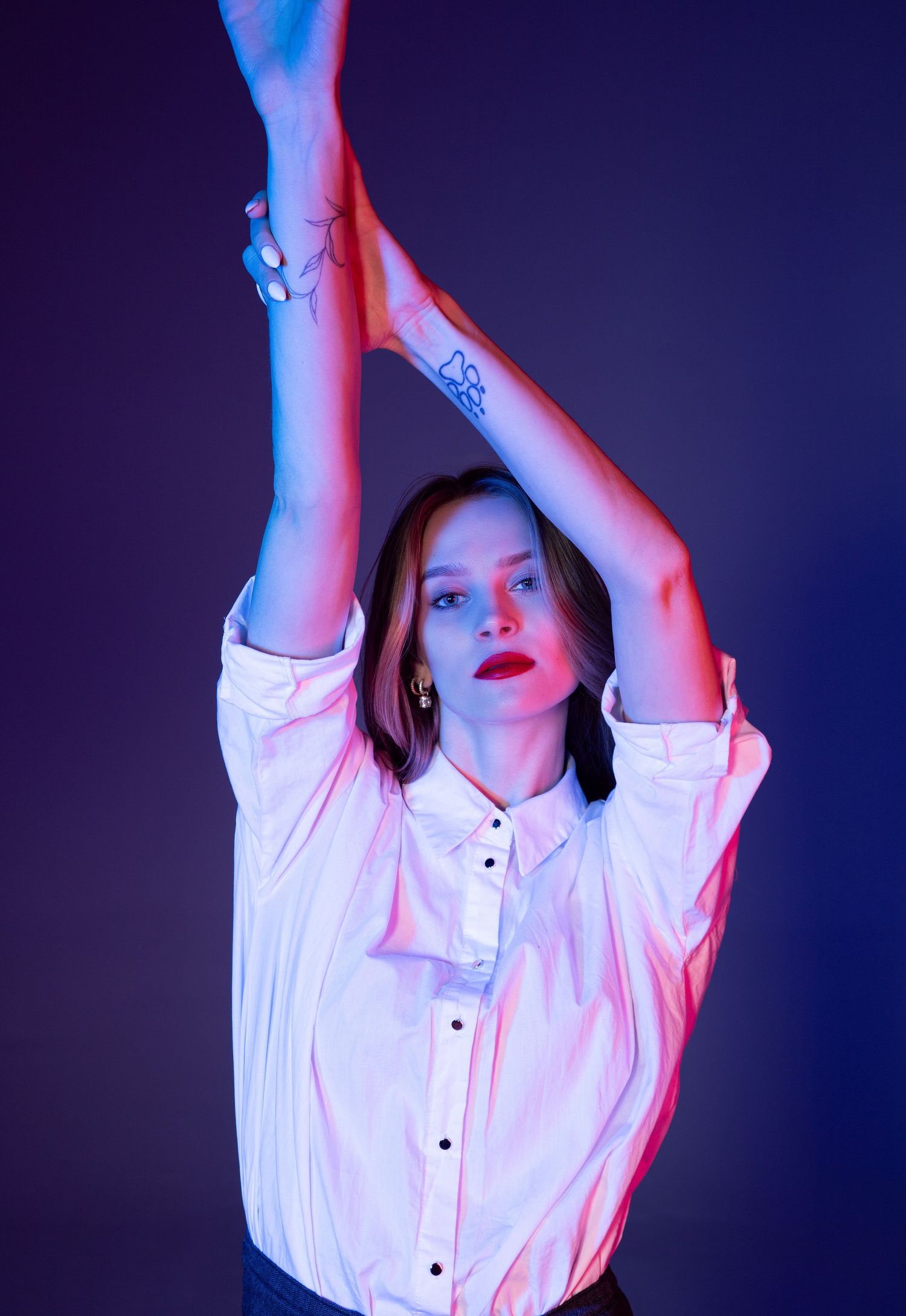 Woman in white shirt raising arms with tattoos visible under blue and pink neon studio lighting.