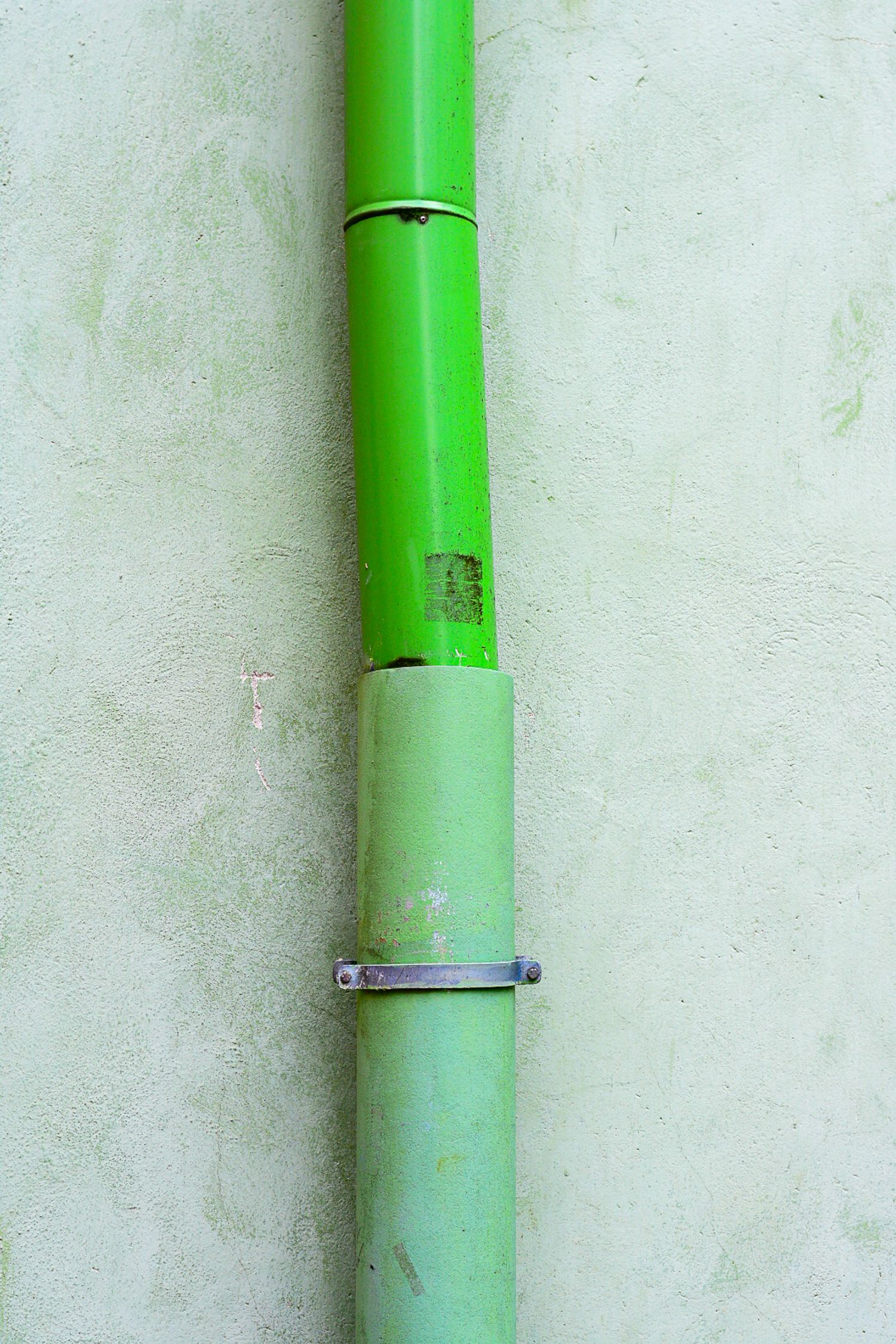 Green painted metal pipe mounted on a light gray concrete wall with a metal bracket clamp securing the joint.