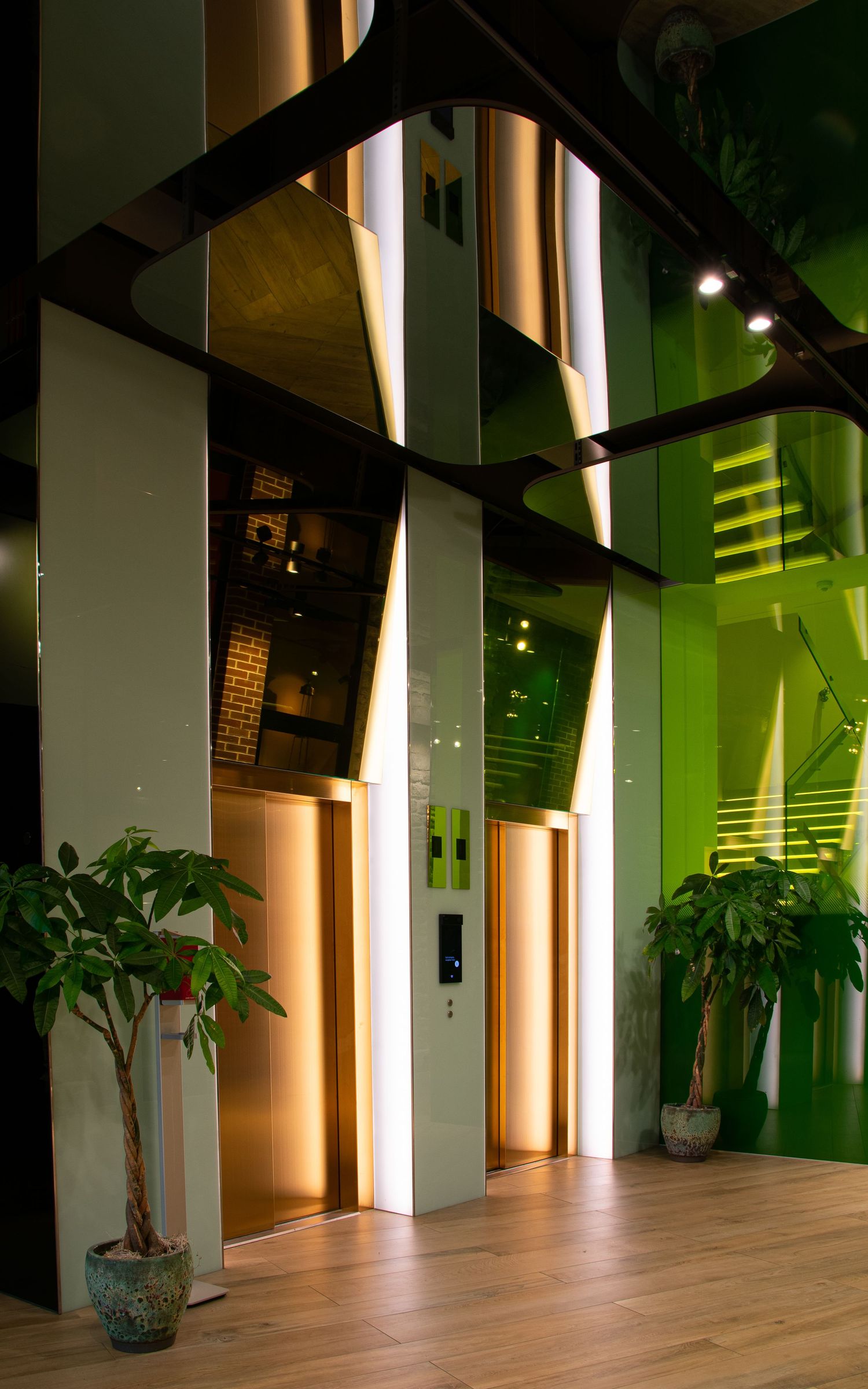 Modern elevator lobby with green glass walls, warm wood panels, and tropical potted plants on a light hardwood floor.