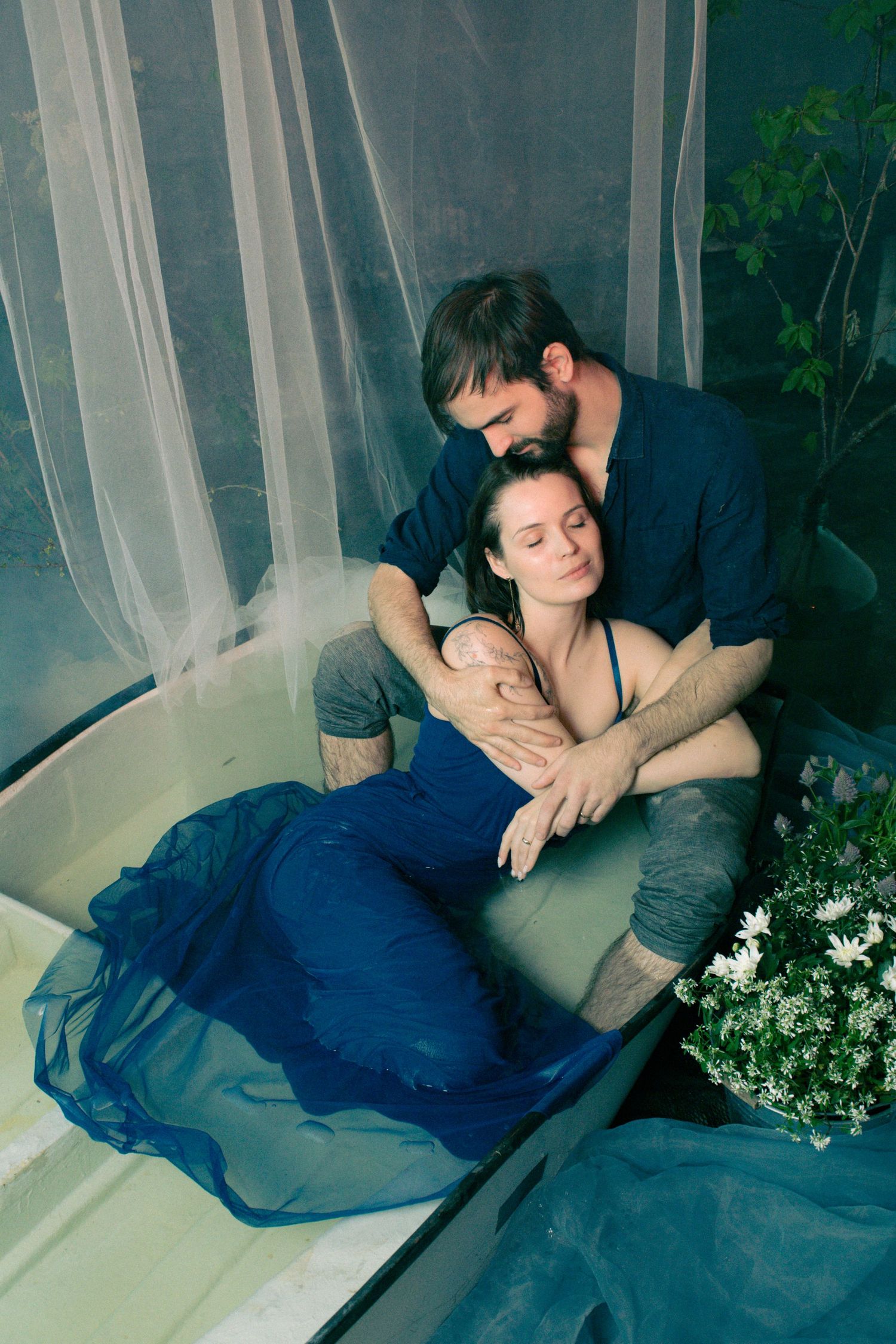 A bearded man tenderly embraces a woman in a blue dress sitting in a bathtub surrounded by white flowers and sheer curtains.