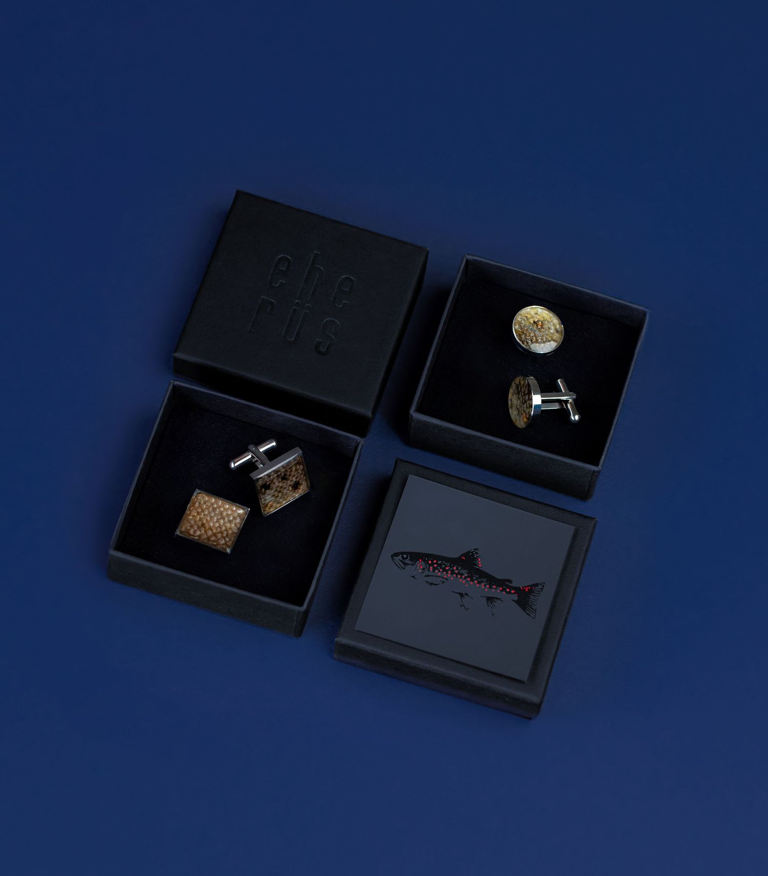 Four luxury cufflinks displayed in open black gift boxes on a dark blue background, featuring gold and silver designs.