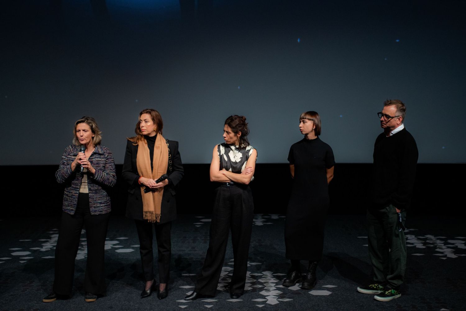 Five people stand on a dimly lit cinema stage in front of a large screen during a film festival Q&A session.