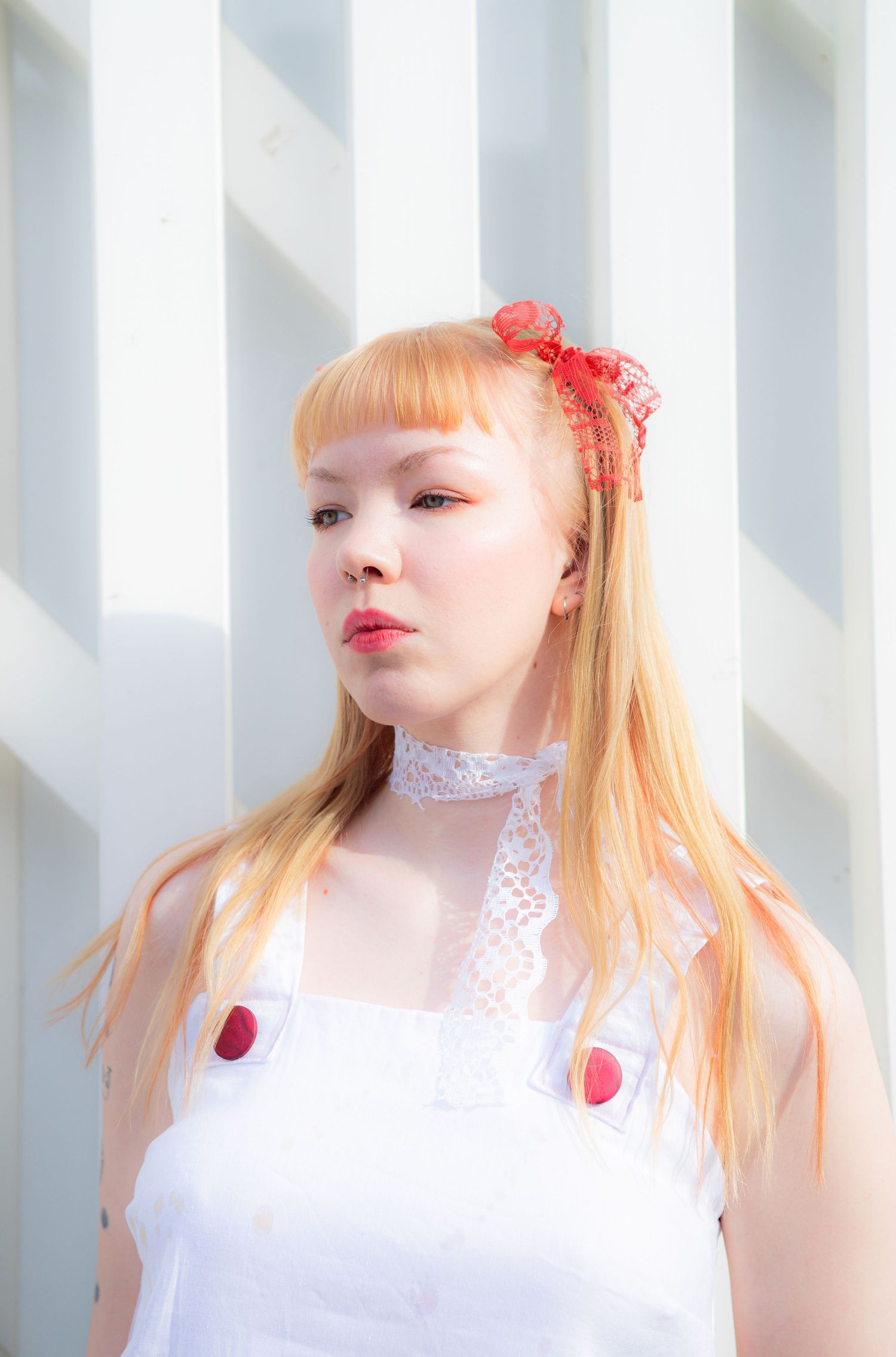 Young woman with orange-blonde hair wearing white dress and red bow hair clip against white wall.