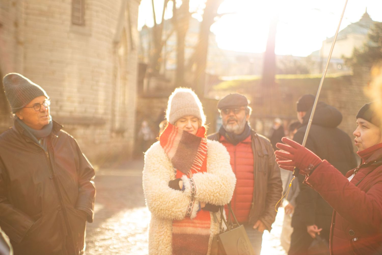 A group of people gather outdoors in golden sunlight near a historic stone building in winter.