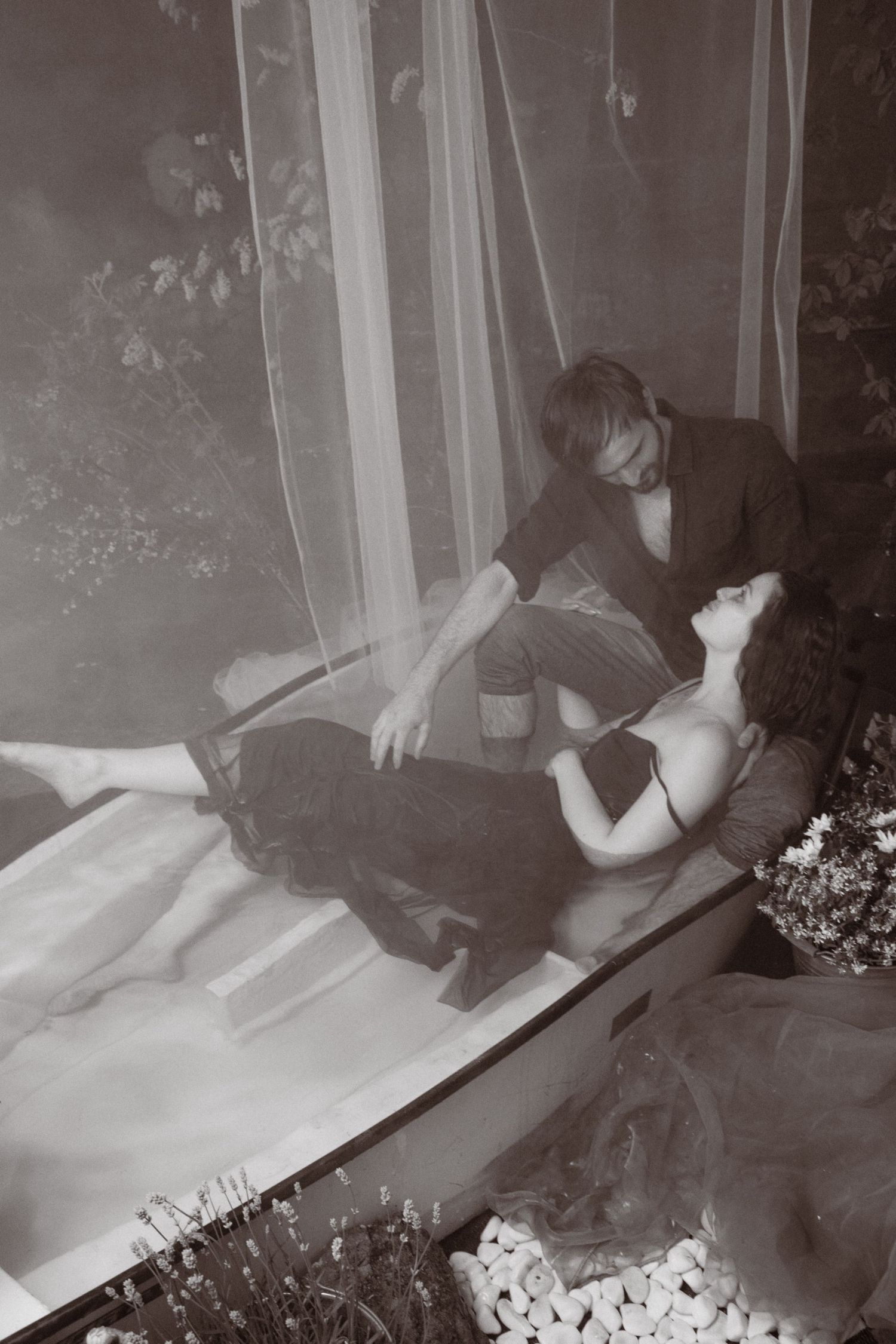 A romantic black and white photo of a couple, woman reclining as man gazes at her near sheer curtains and flowers.