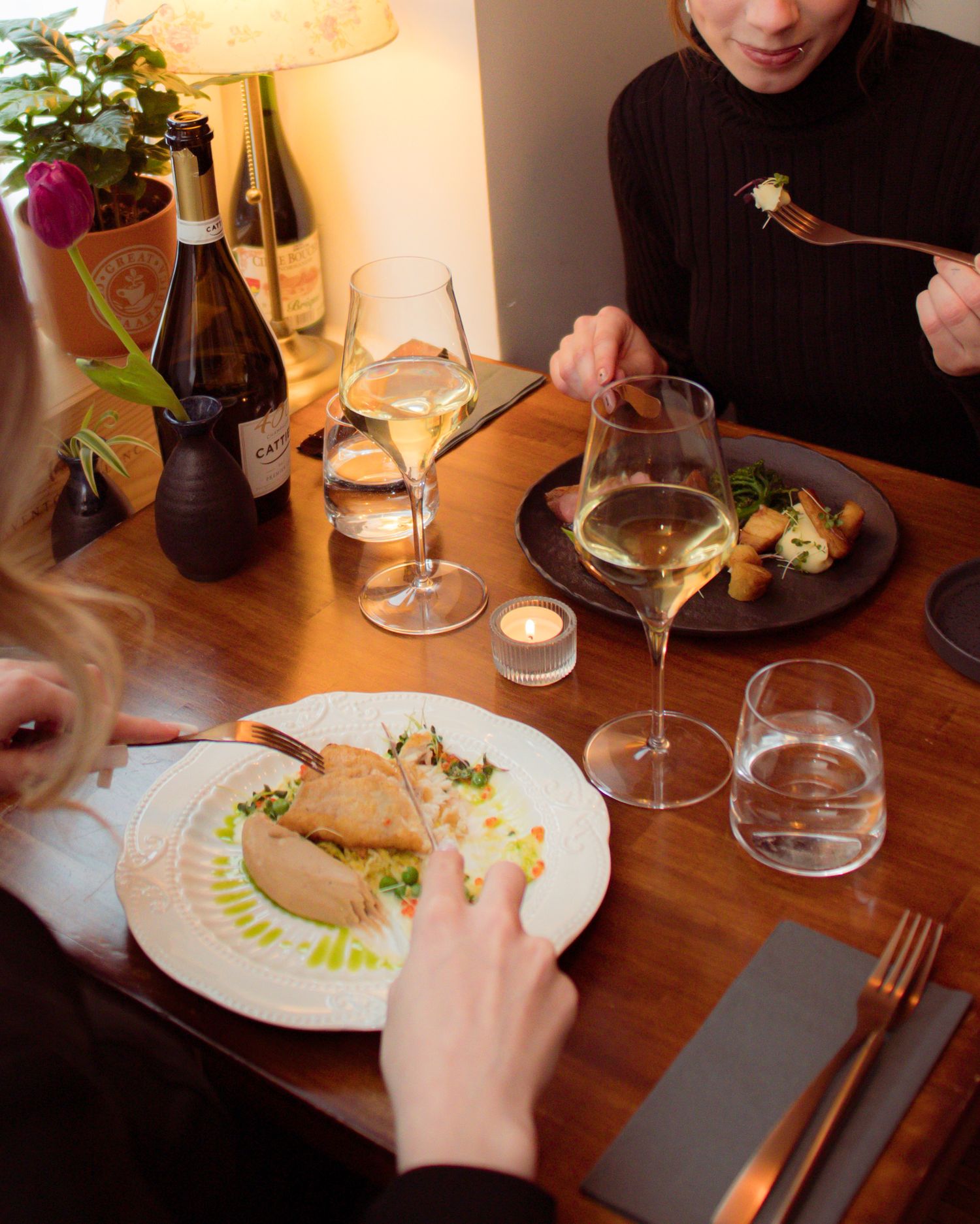 Two women dining at a wooden table with elegant plated dishes and white wine glasses in a warmly lit restaurant.