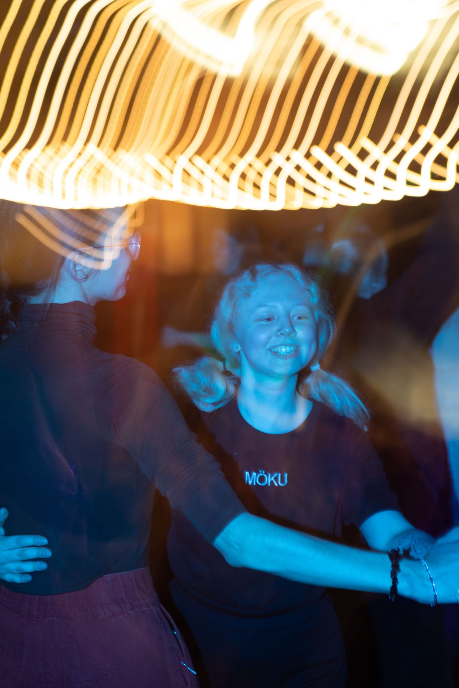 Smiling woman in Moku shirt dancing under streaking golden light trails at a night event.
