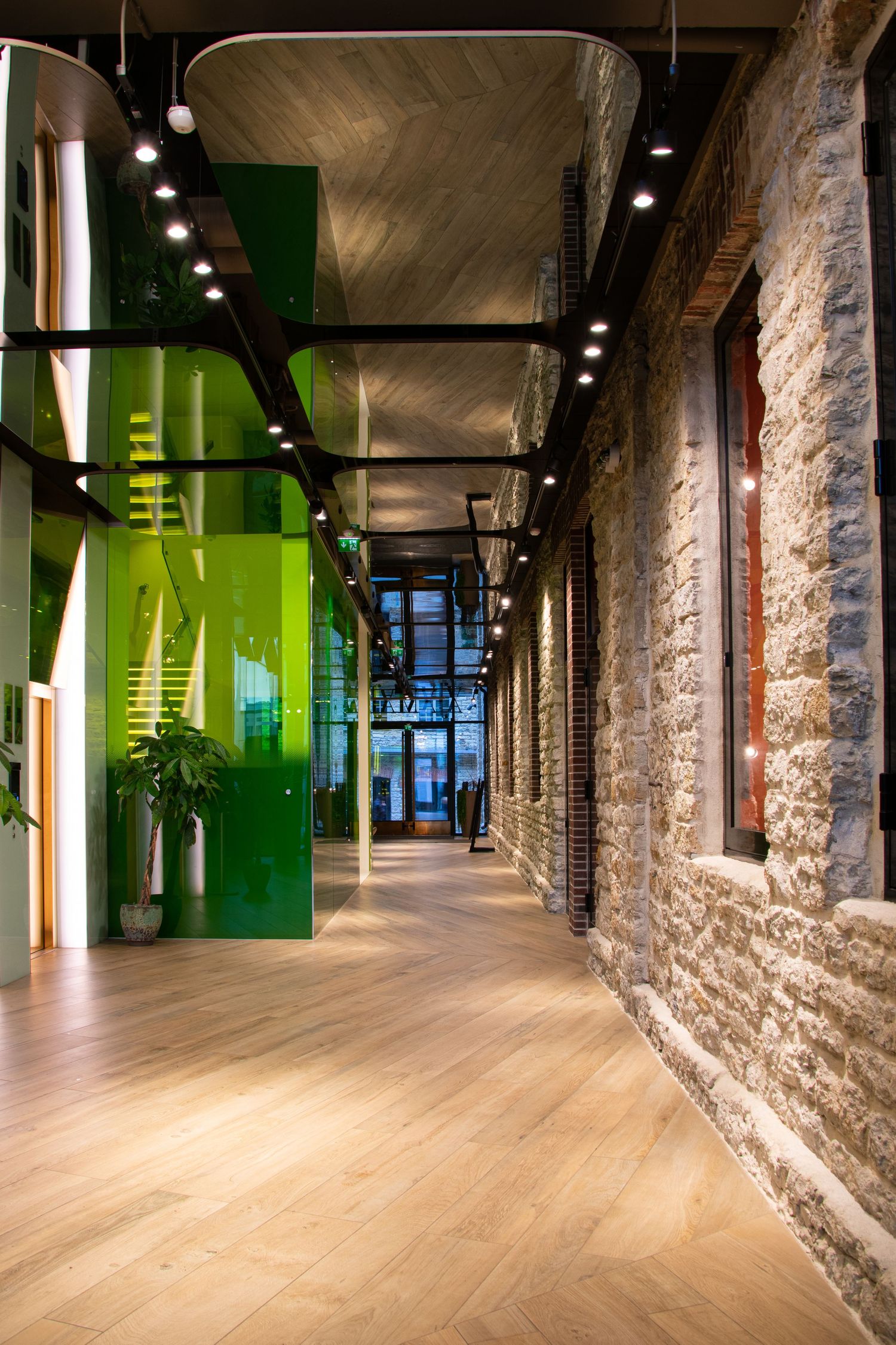 Modern corridor with exposed stone brick wall, green glass partition, wood floor, and industrial track lighting.