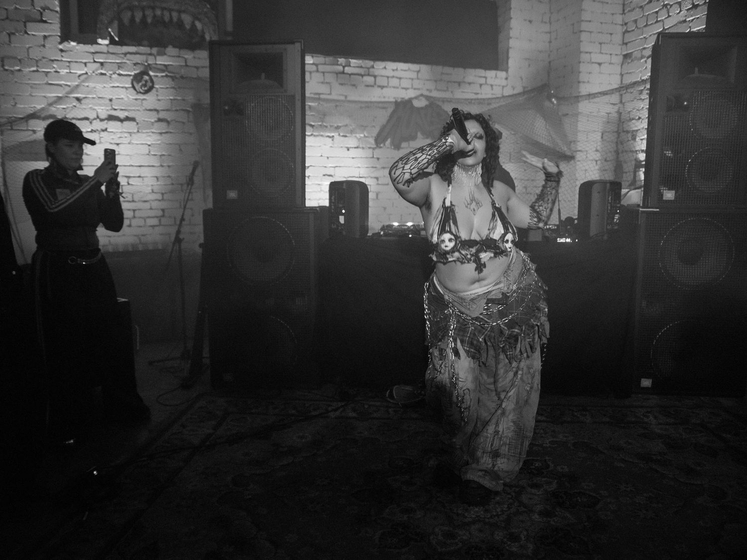 Black and white photo of a belly dancer performing on stage in a dimly lit brick venue.