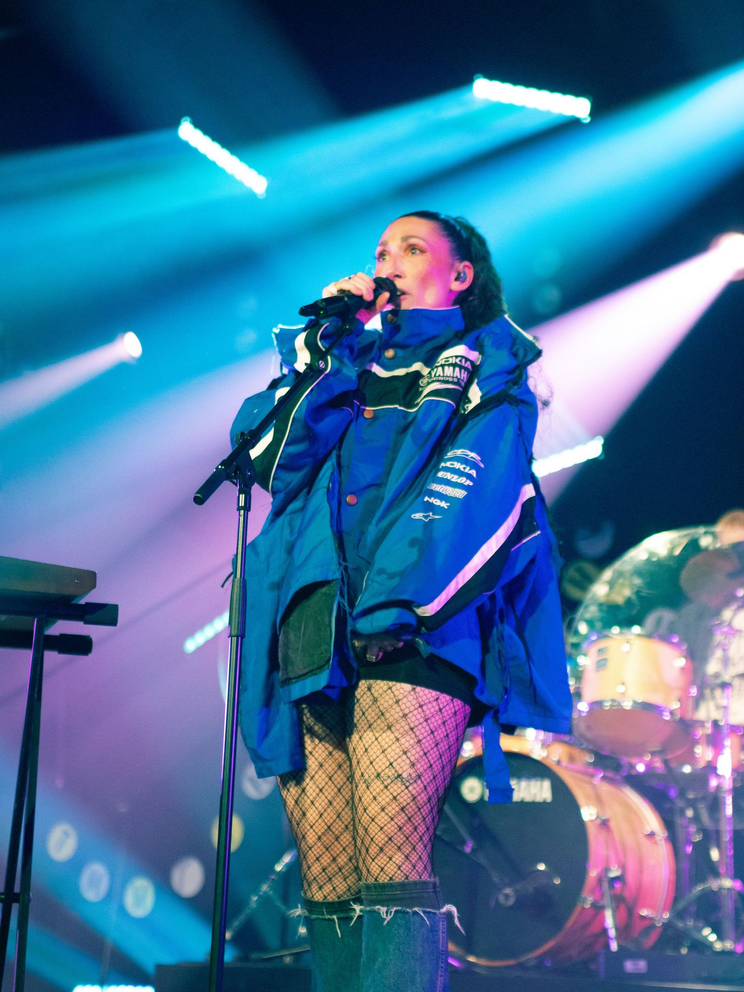 Female vocalist singing on stage in a blue Yamaha jacket with fishnet stockings and green boots.