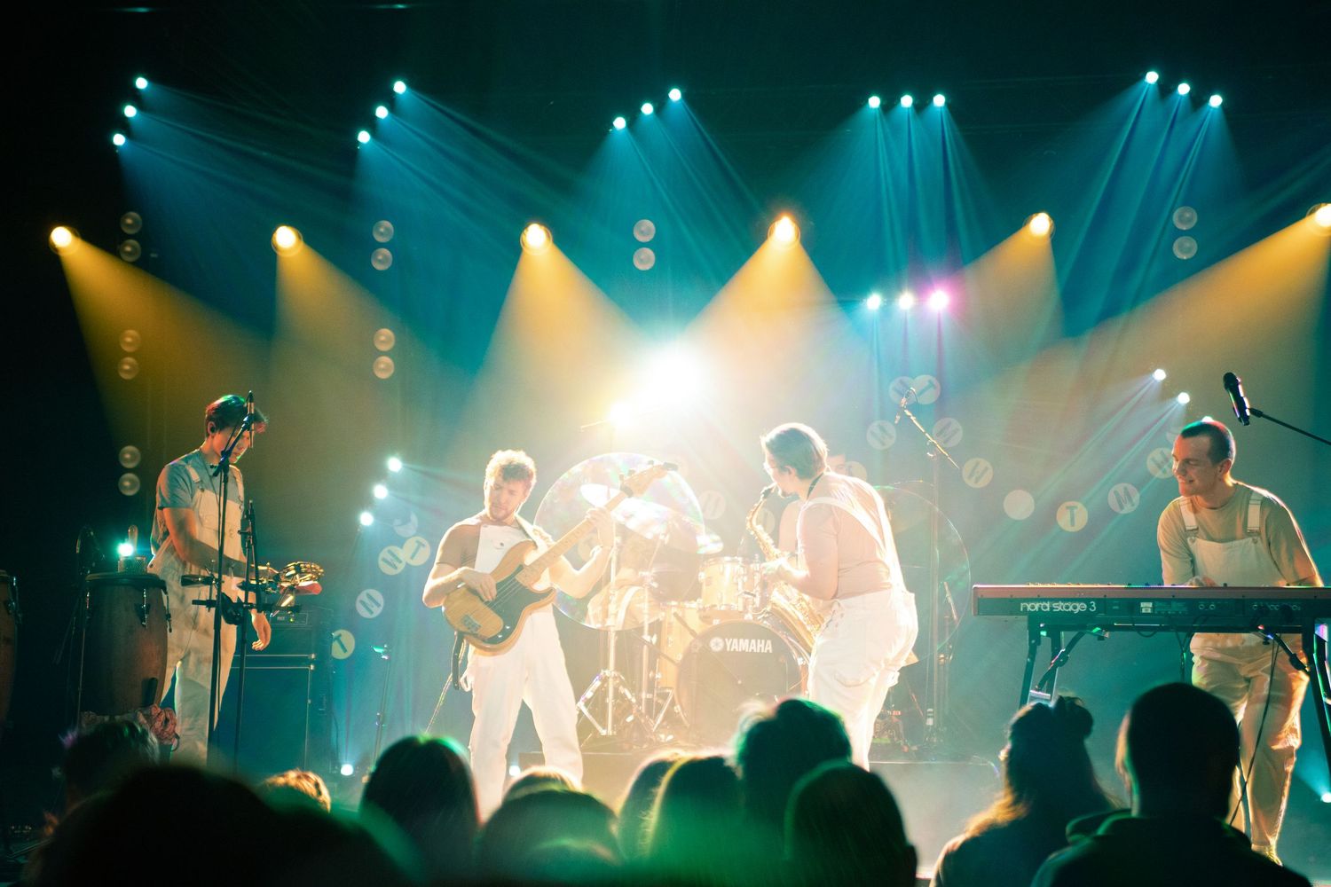 Full band performing on a brightly lit stage in white outfits before a live audience.