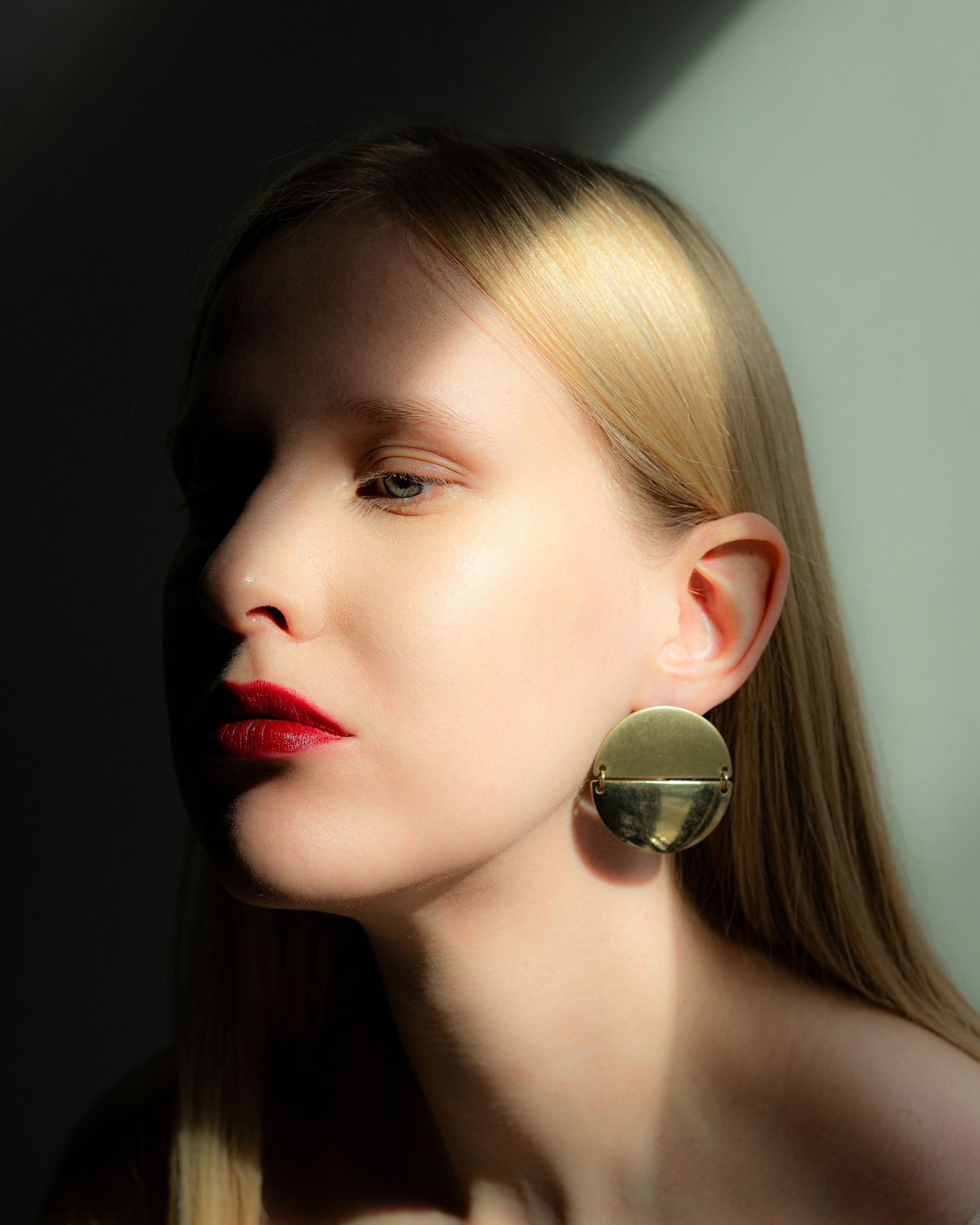 Blonde woman wearing bold gold circular earrings with red lips in a dramatic shadow portrait.