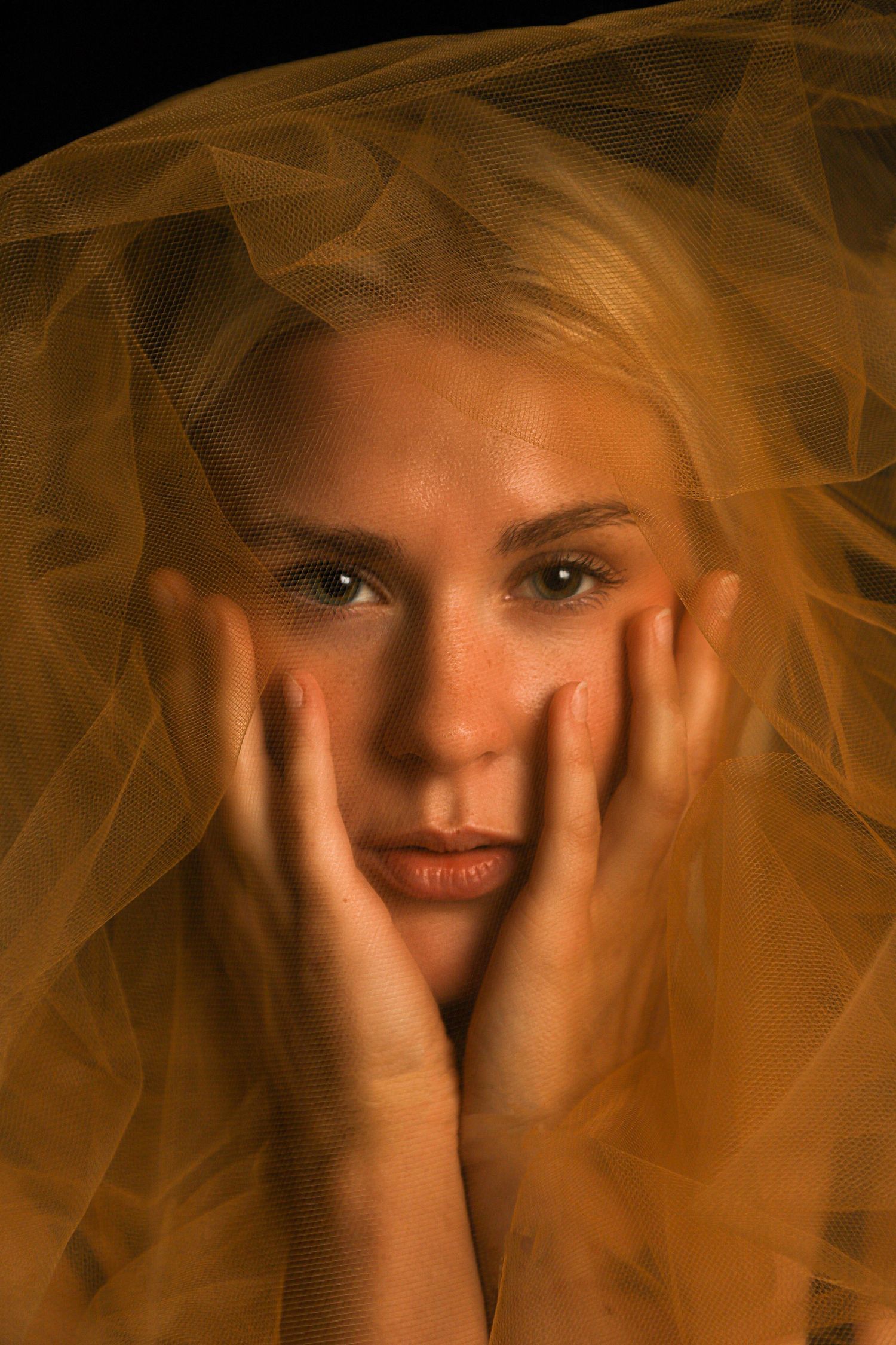 Blonde woman with hands on cheeks covered by golden yellow tulle veil against dark black background.