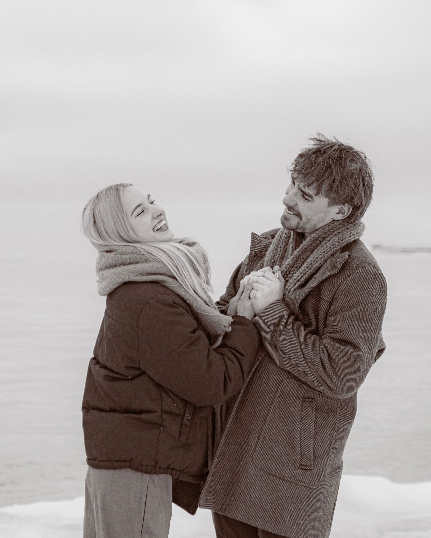 Black and white photo of a joyful couple holding hands in winter coats near a snowy waterfront.