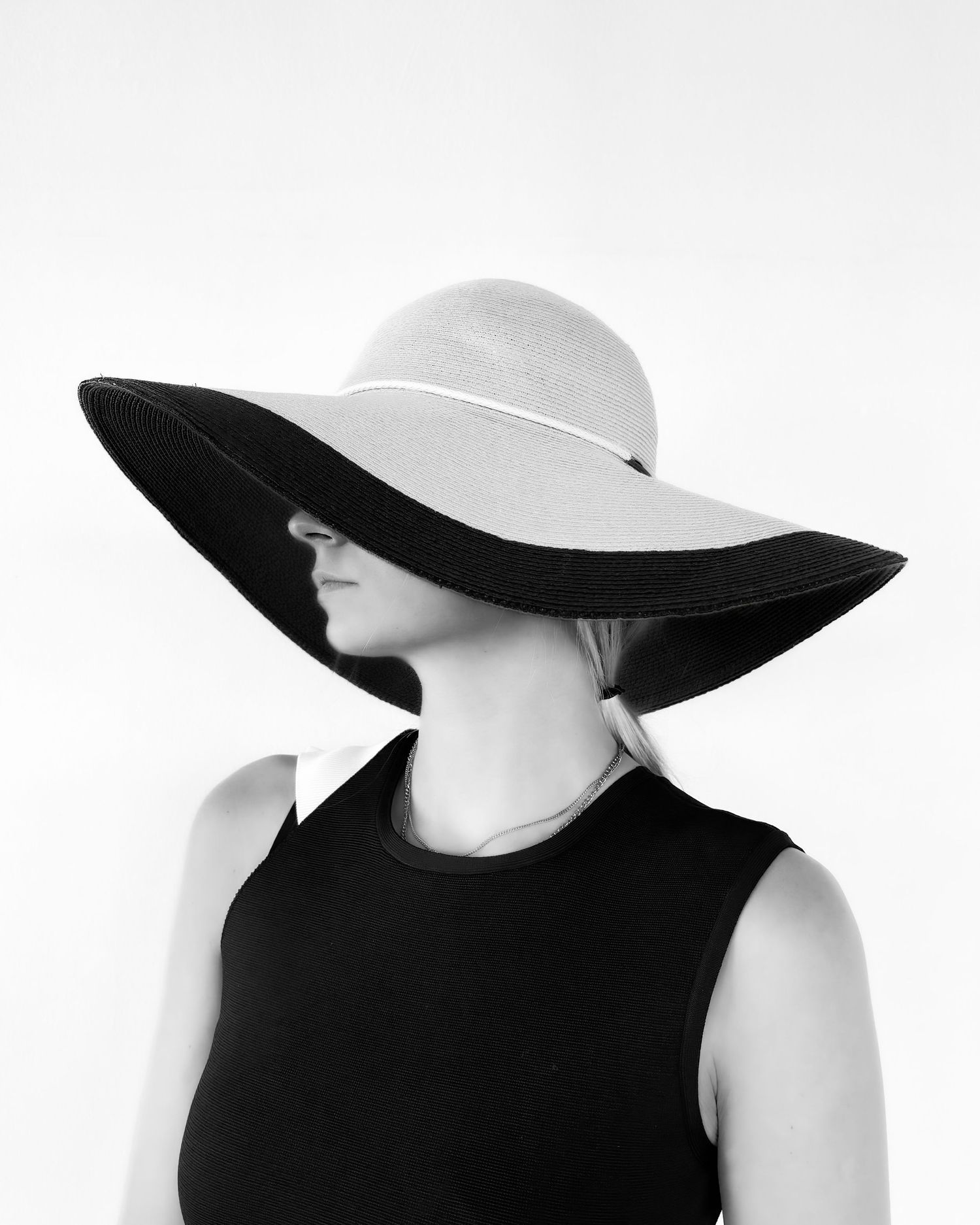 Black and white photo of a woman in a black dress wearing a wide-brim floppy sun hat covering her face.