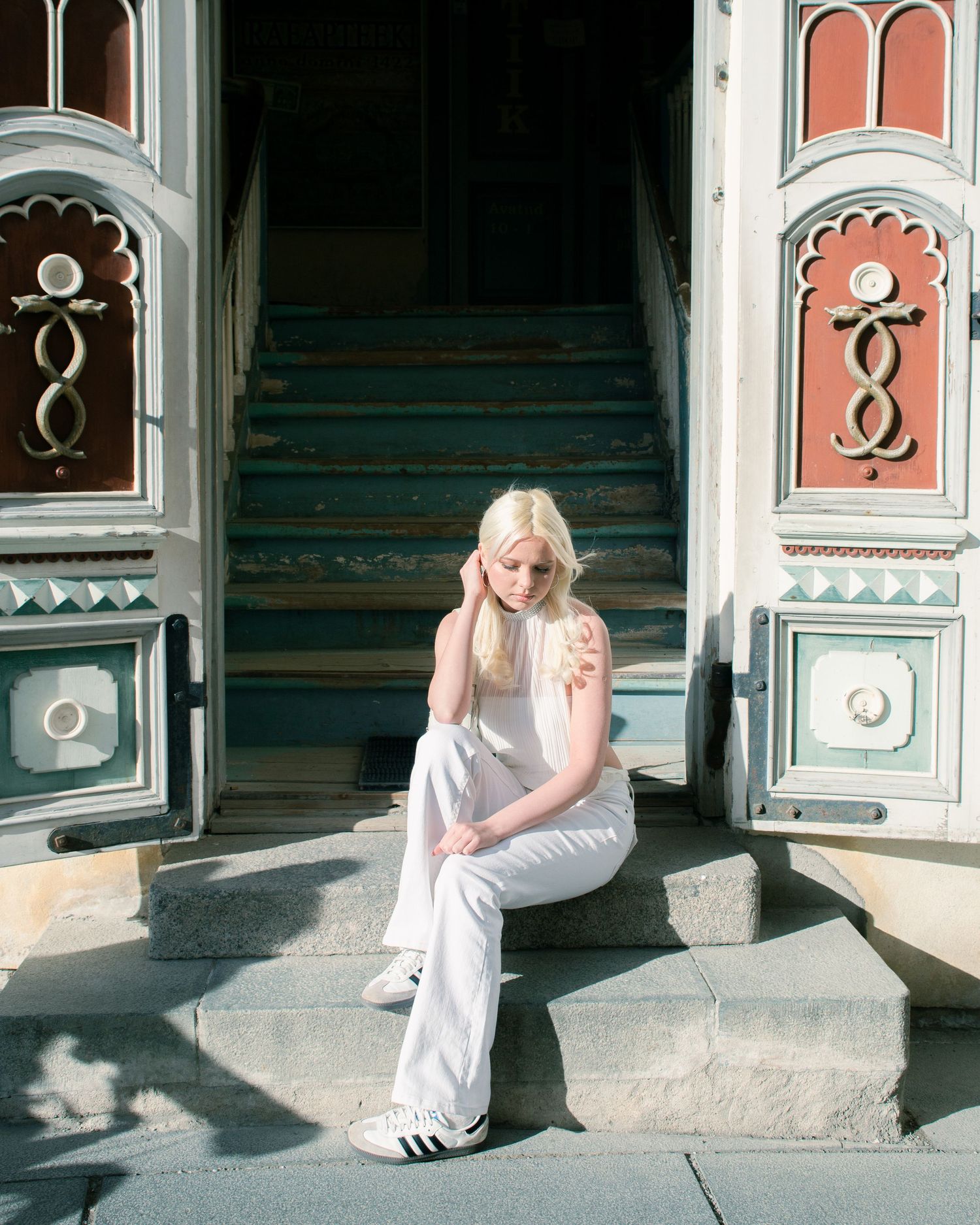 Blonde woman in white outfit sitting on ornate building steps in sunlight.