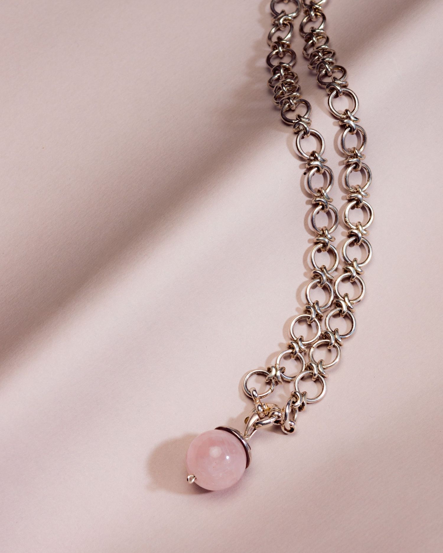 Silver chainmail necklace with a pink rose quartz ball pendant on a soft pink fabric background.