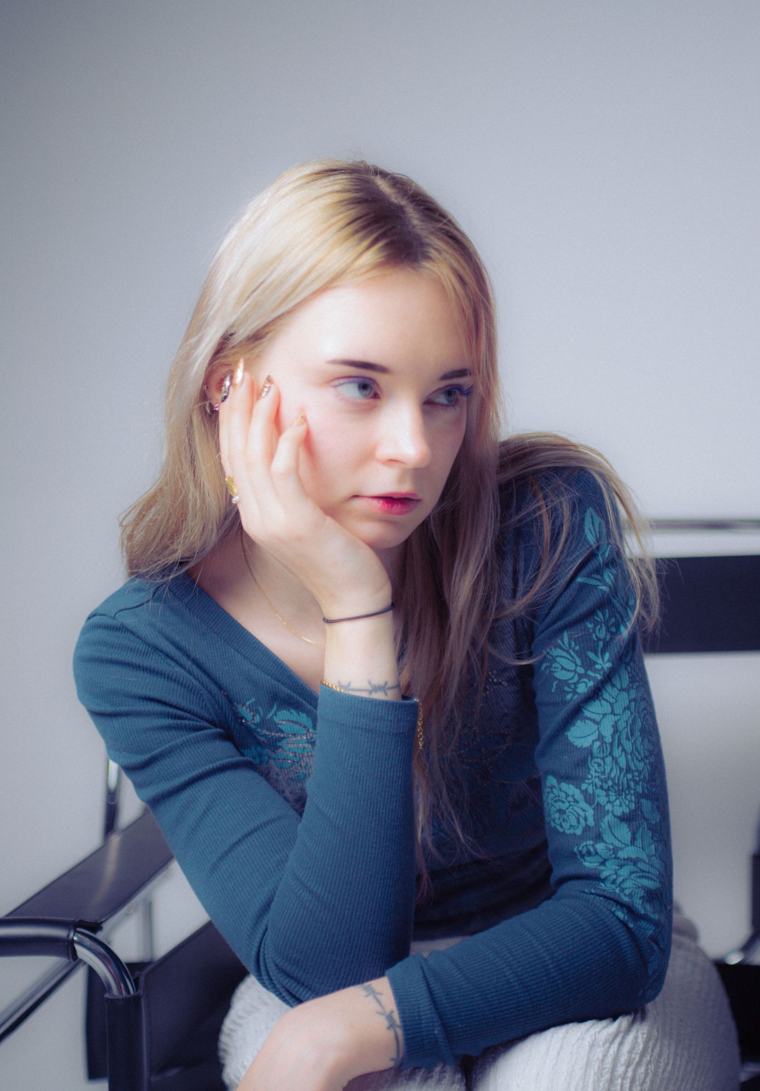 A young blonde woman in a teal floral top resting her chin on her hand while seated indoors.