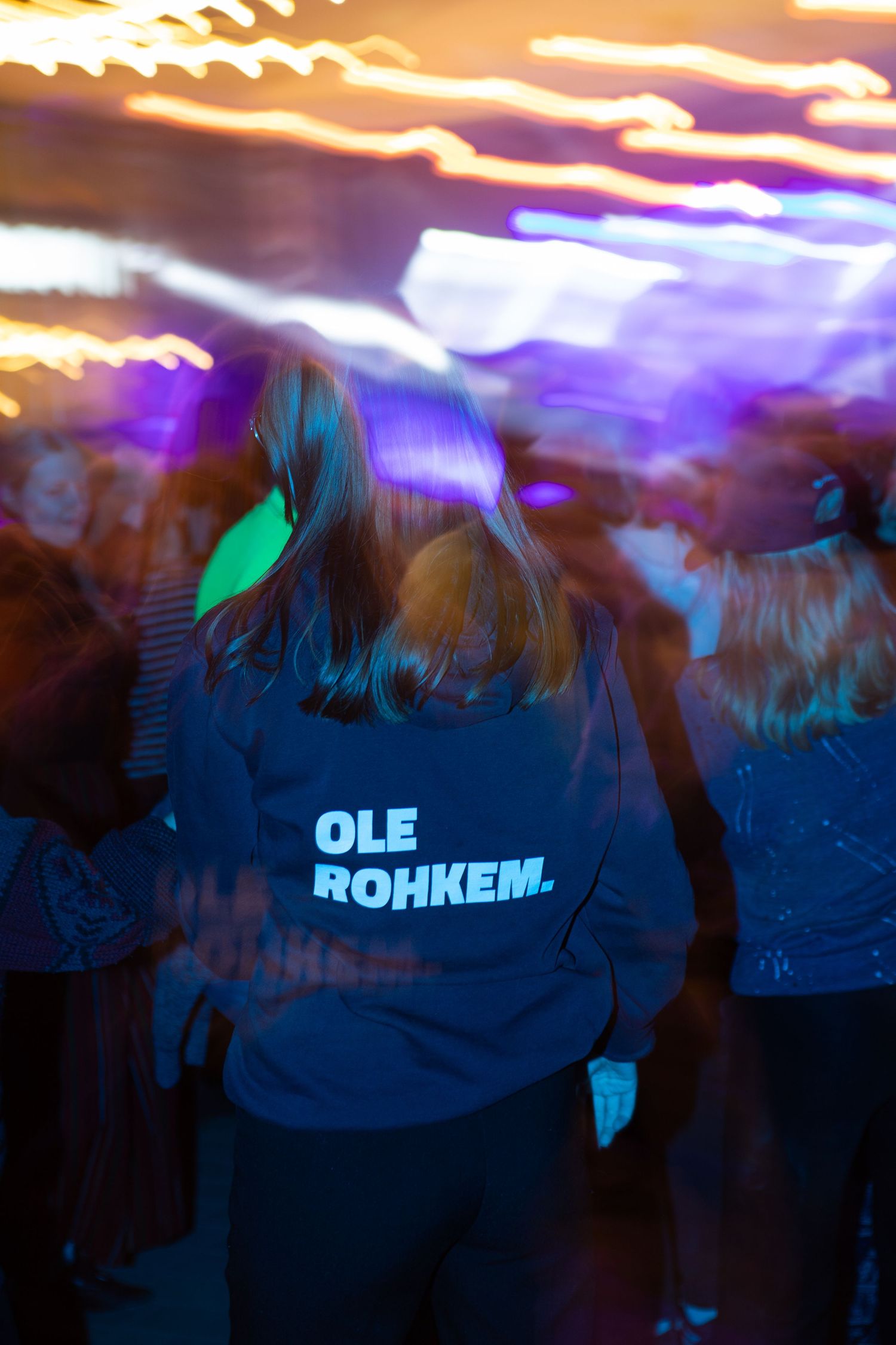 Person wearing Ole Rohkem hoodie standing in a crowd at a colorful outdoor night concert.